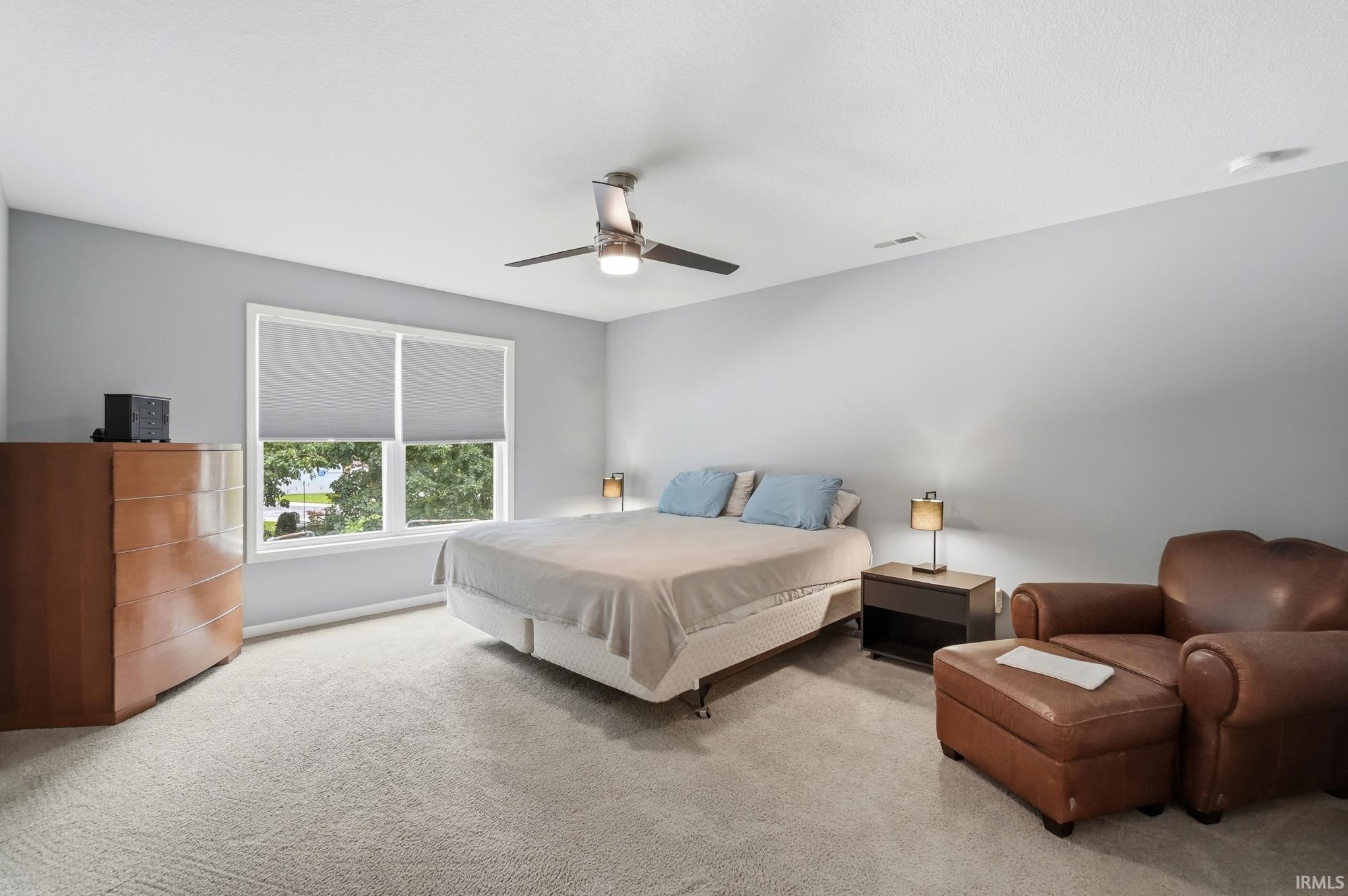 Bright bedroom featuring a large window, neutral carpeting, a ceiling fan with integrated lighting, and light gray wall paint