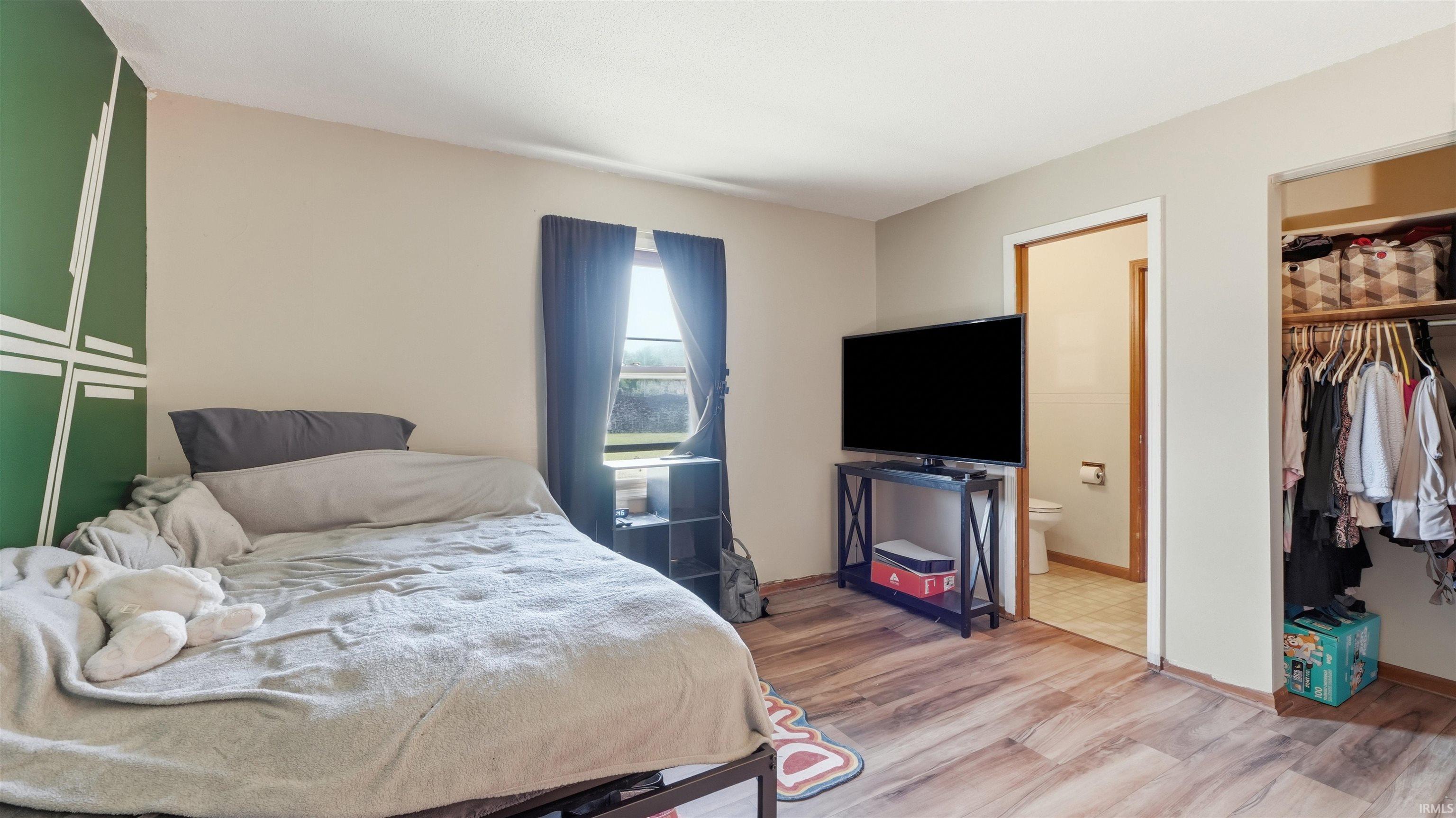 Bedroom featuring wood-finish flooring, a decorative accent wall, a window with dark curtains, an open doorway, and an open closet