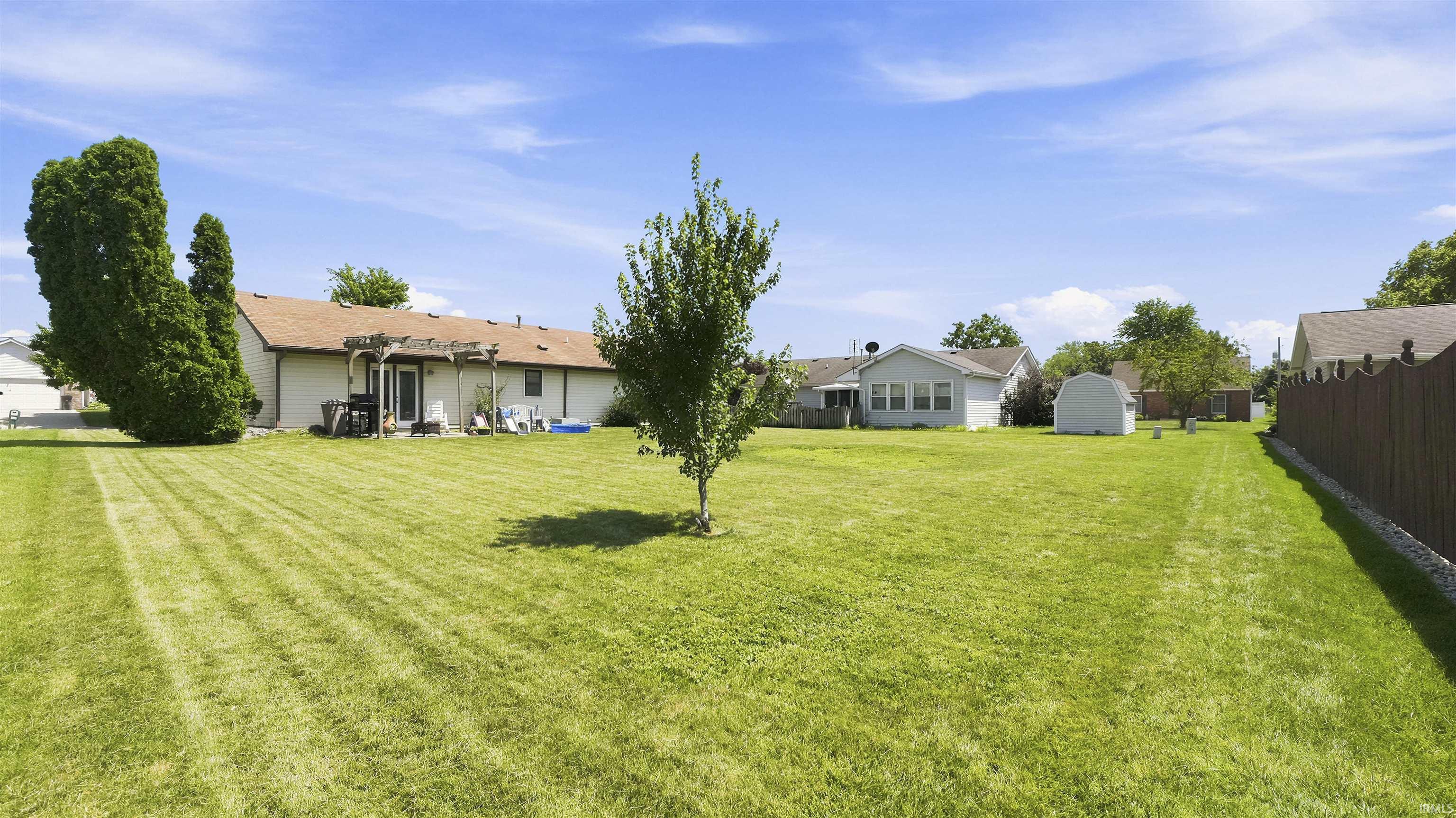 Expansive grassy backyard featuring a perimeter privacy fence, mature trees, a built-in outdoor structure, and a storage shed