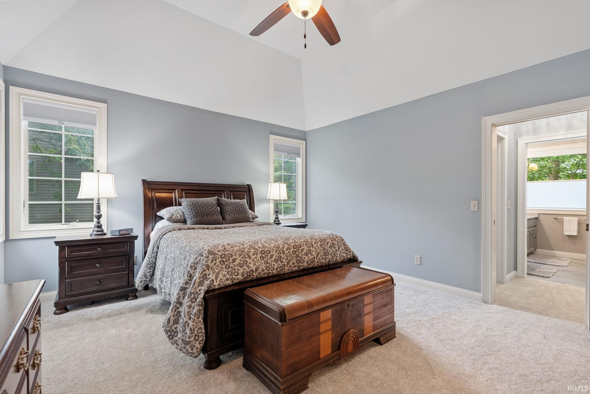 Carpeted sleeping area featuring a vaulted ceiling with a ceiling fan, dual windows, and an entryway to an adjoining bathroom with a window