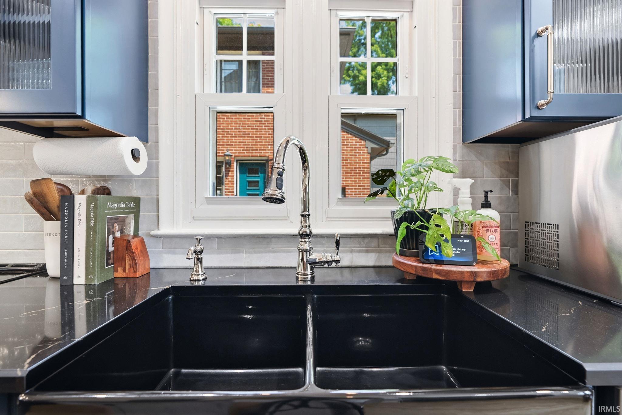 Kitchen sink featuring a dual-basin black farmhouse style sink, polished chrome gooseneck faucet, black countertop, light grey subway tile backsplash, and blue cabinetry with fluted glass accents