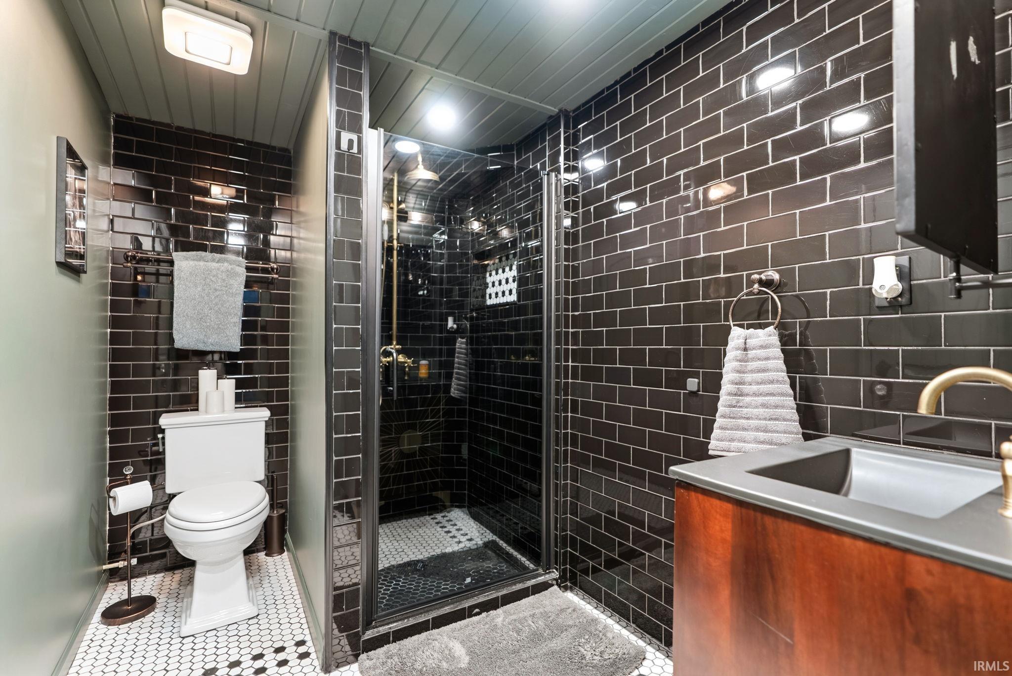 Bathroom featuring black subway tile walls, a glass-enclosed shower with black and white hexagon tile flooring, a wood-finish vanity with an integrated sink and brass-tone faucet, and white hexagon tile flooring