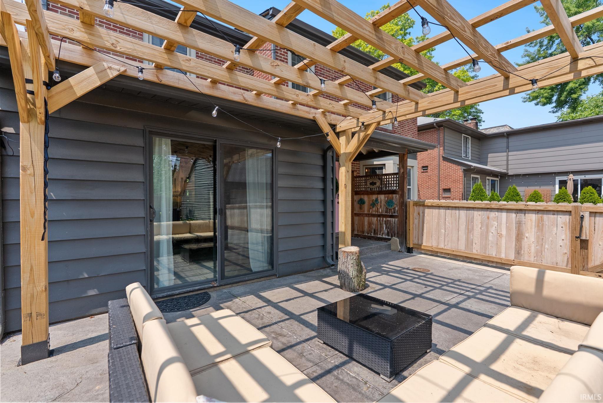 Expansive outdoor patio featuring a wooden pergola, sliding glass door entry, grey horizontal siding, concrete pavers, and a wood privacy fence