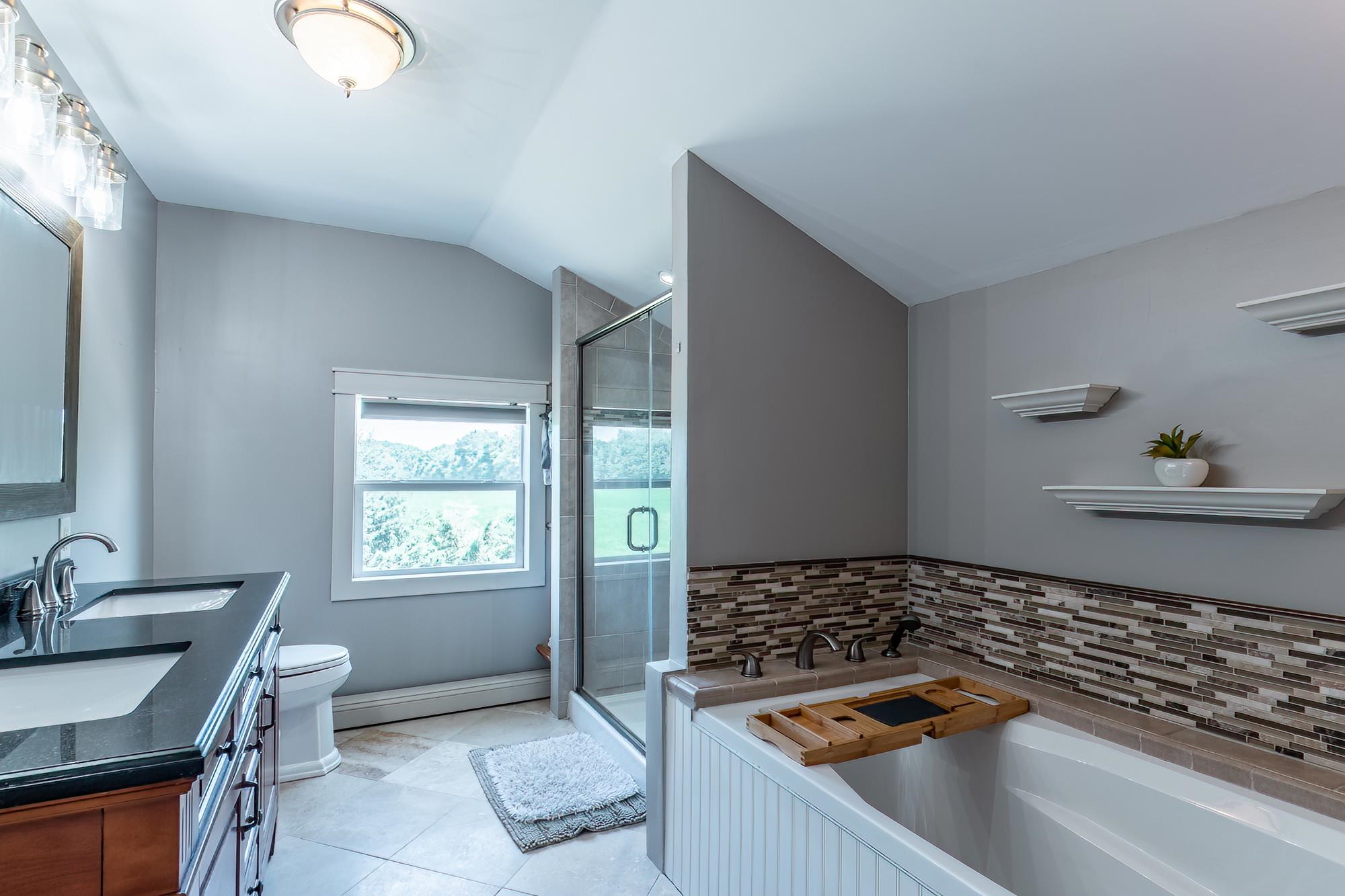 Bathroom featuring a dual vanity with dark countertops, a built-in tub with a mosaic tile surround, a glass-enclosed shower, and a white window with exterior visibility