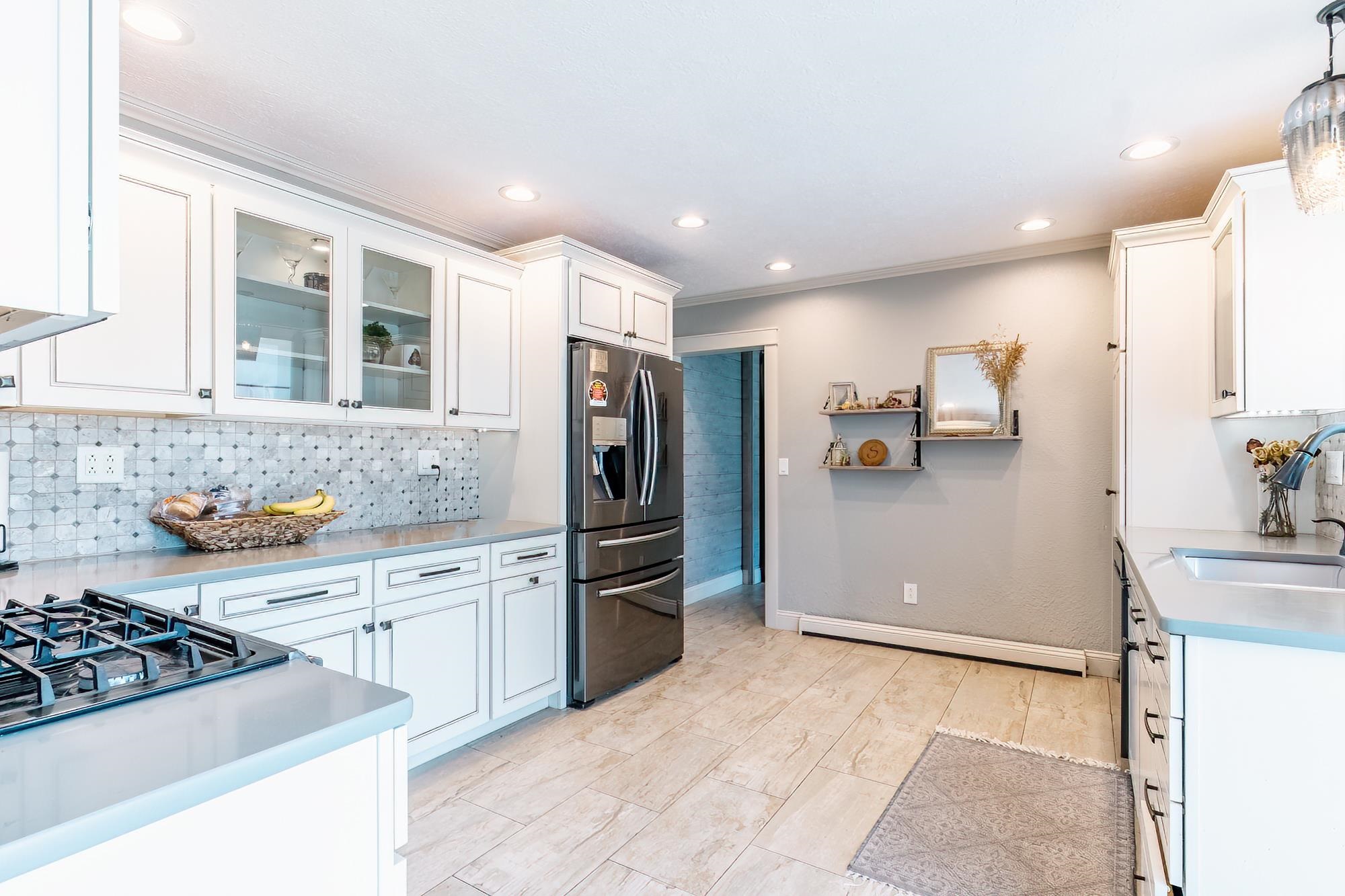 White shaker cabinetry with glass-front accents, dark stainless steel appliances, a gas cooktop, patterned tile backsplash, and light-toned tile flooring