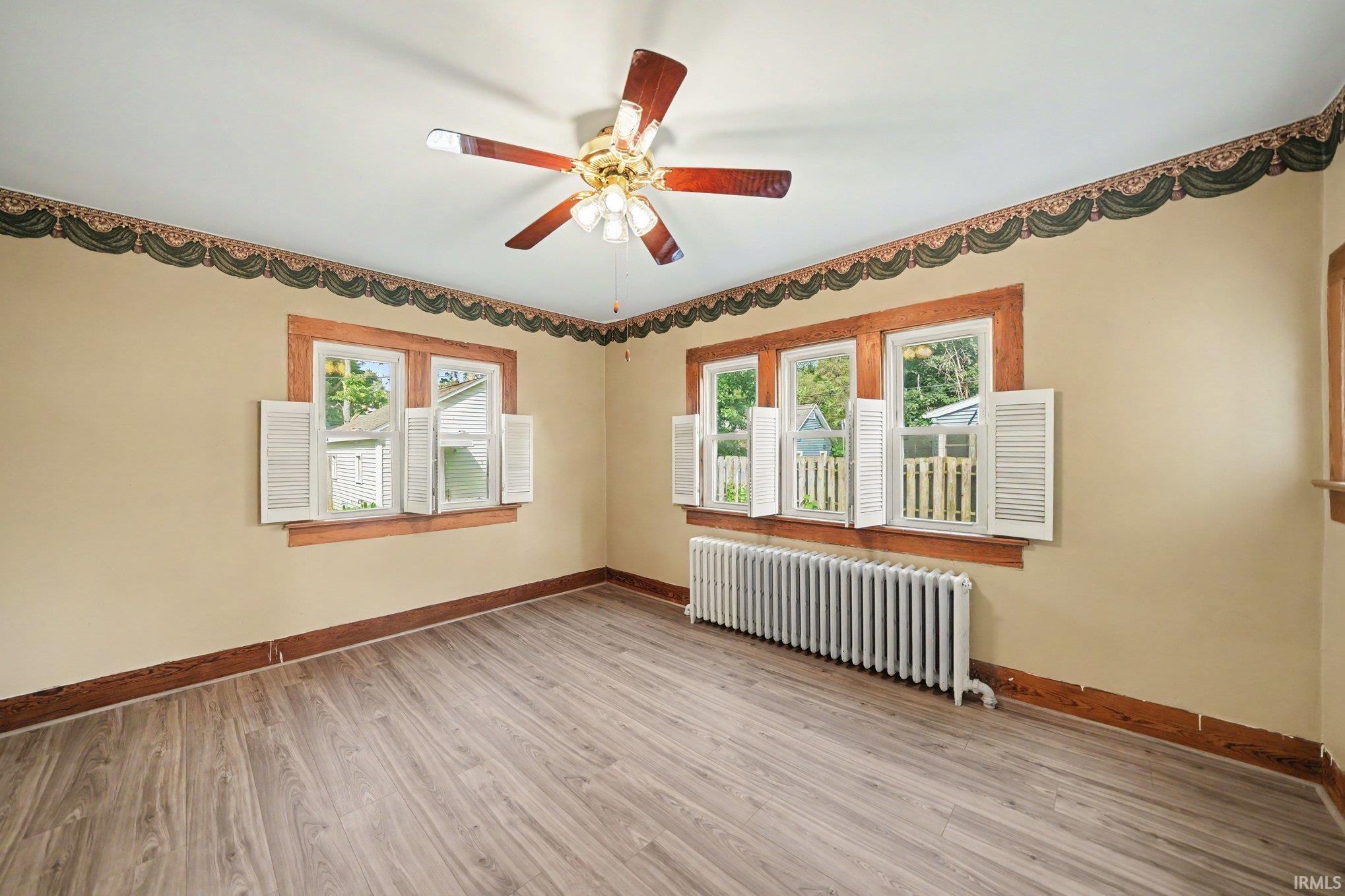 Spacious room featuring wood-finish flooring, two windows with white louvered shutters, and a multi-light ceiling fan