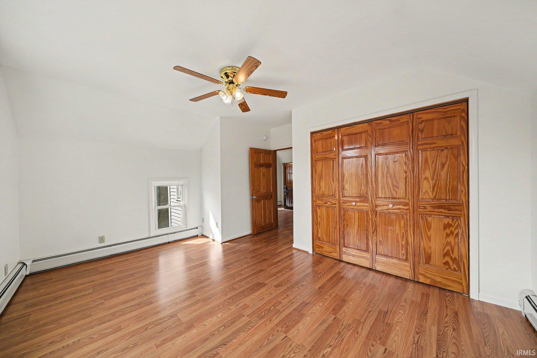 Wood-finish flooring throughout, featuring a multi-panel closet with bi-fold doors, a ceiling fan with integrated lighting, and white baseboard heating