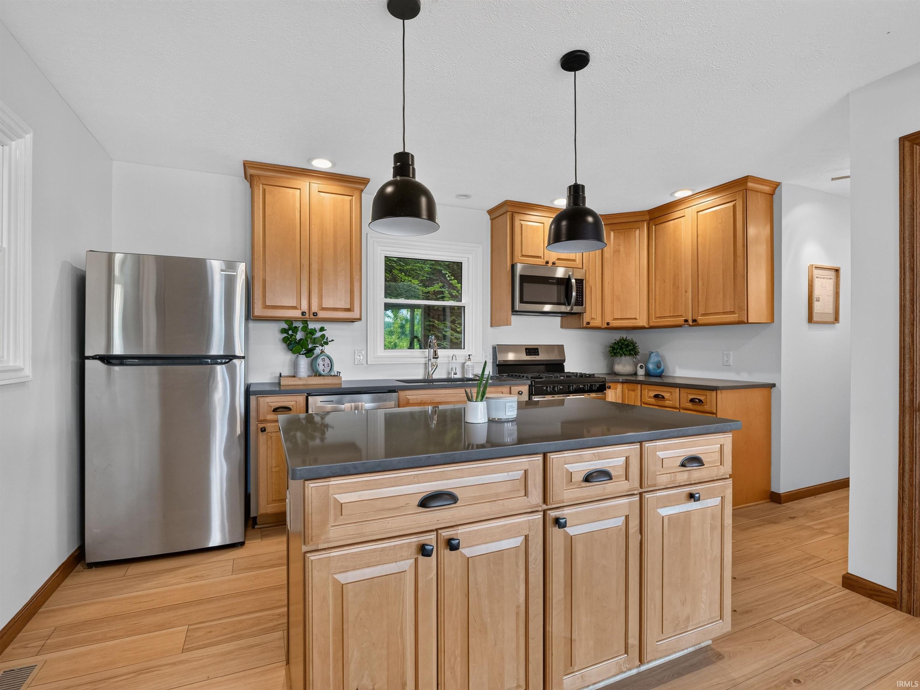 Kitchen featuring a central island with storage, wood-finish flooring, stainless steel appliances, light wood cabinetry, and dark countertops