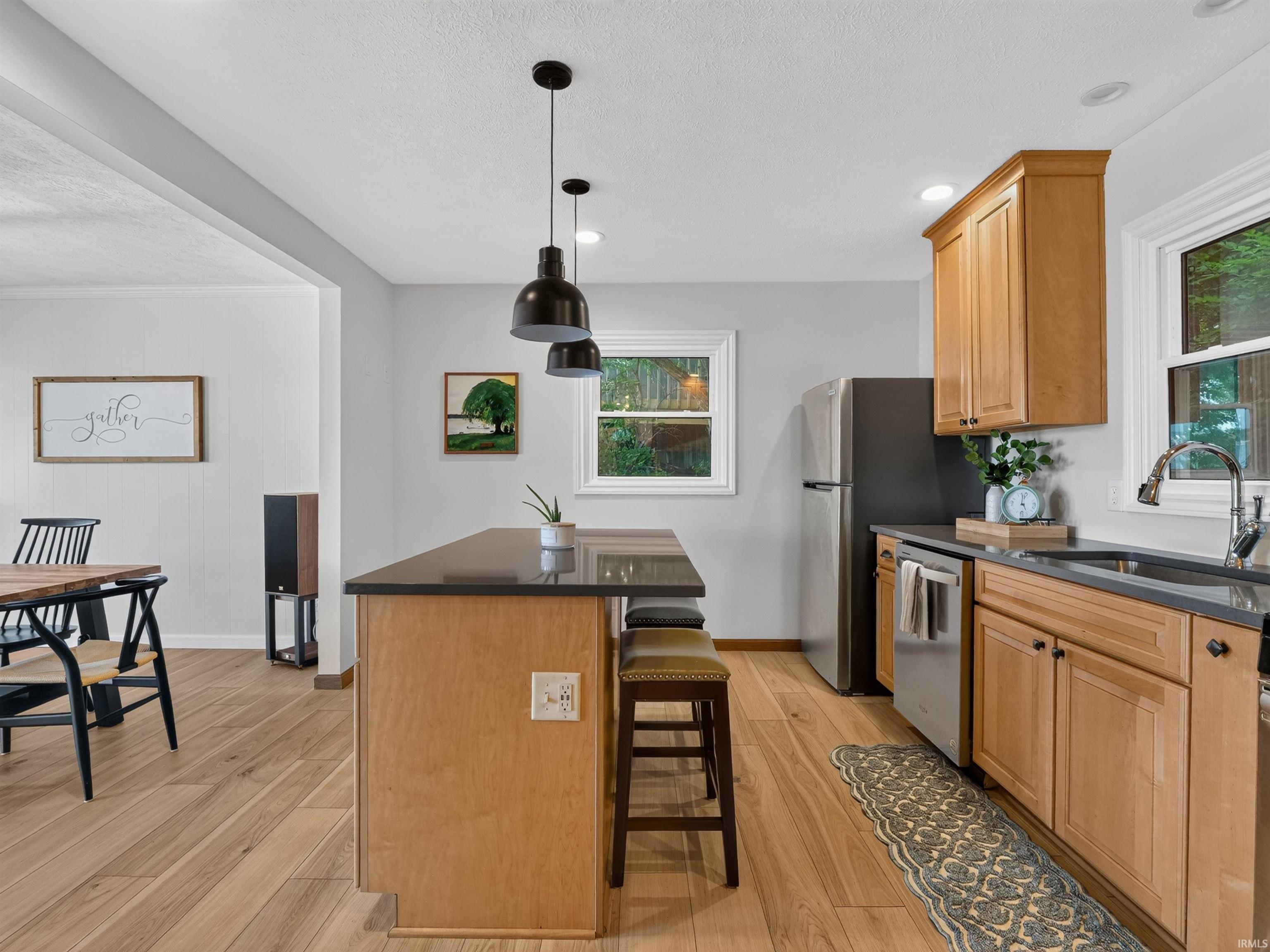 Kitchen featuring light wood-finish flooring, a dark countertop island with pendant lighting, light wood cabinetry, and stainless steel appliances