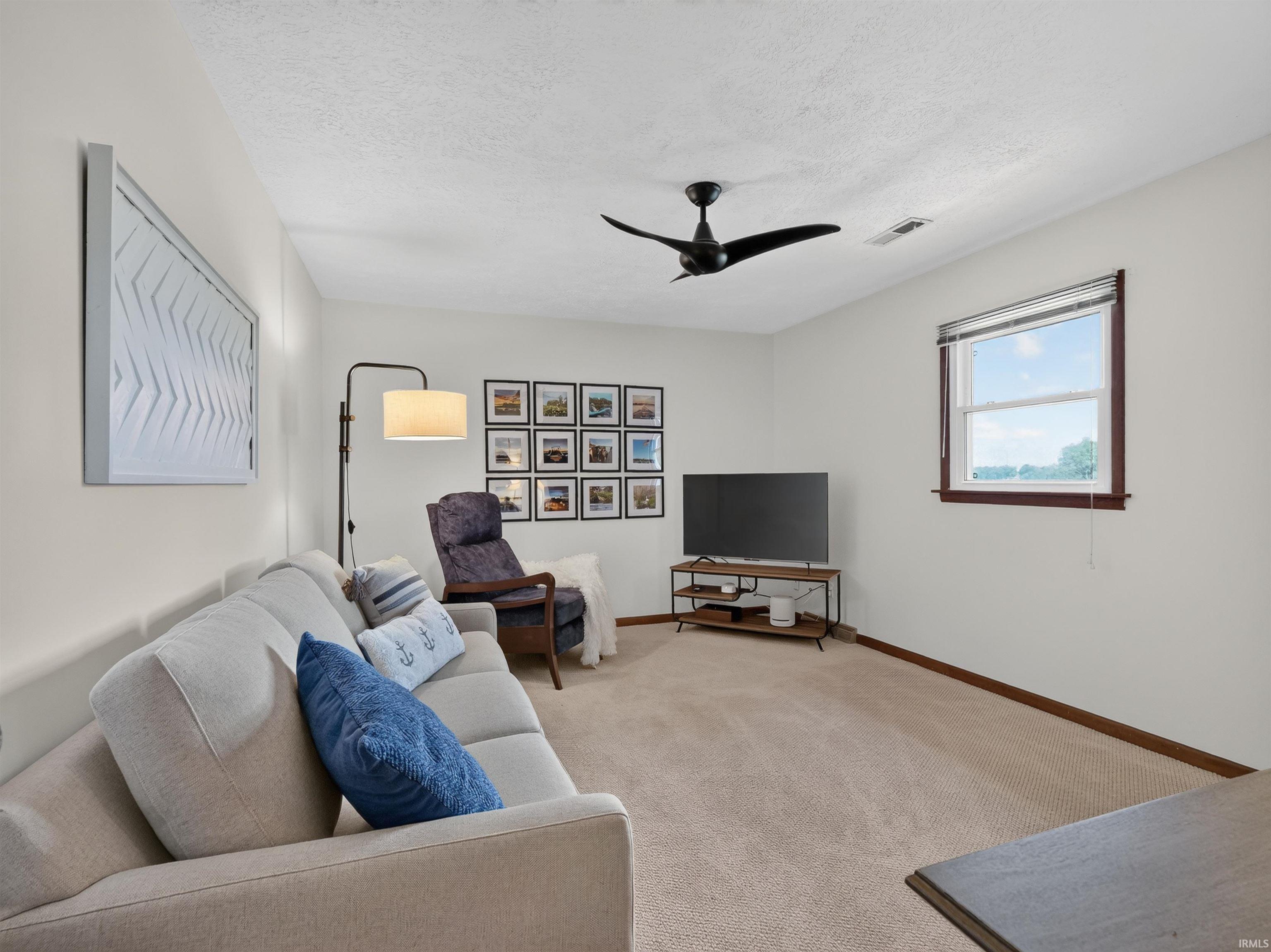 Carpeted living area featuring a ceiling fan, white walls, and a window with wood-finish trim