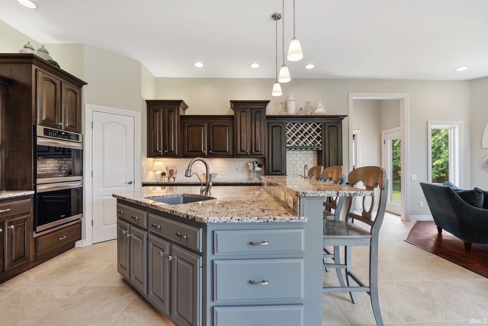 Kitchen featuring a multi-tone island with a built-in sink and granite countertop, dark wood-finish cabinetry, stainless steel built-in double oven, integrated wine rack, and recessed lighting
