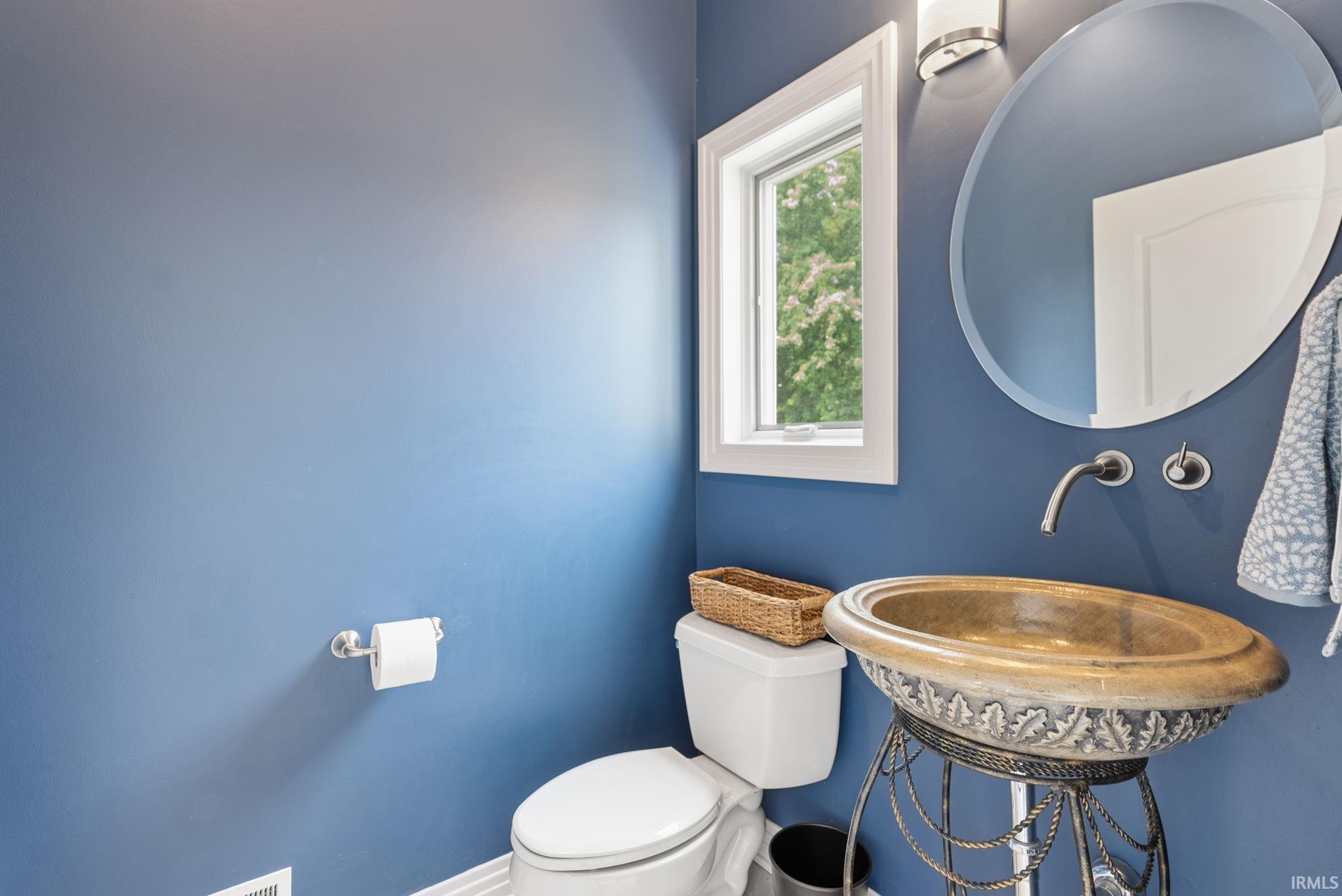Powder room featuring a decorative vessel sink, wall-mounted faucet, circular mirror, and a window with white trim
