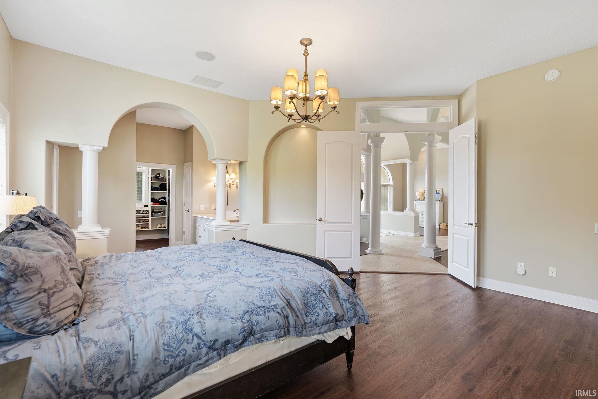 Spacious room featuring dark wood-finish flooring, a decorative chandelier, arched doorways, architectural columns, and built-in shelving