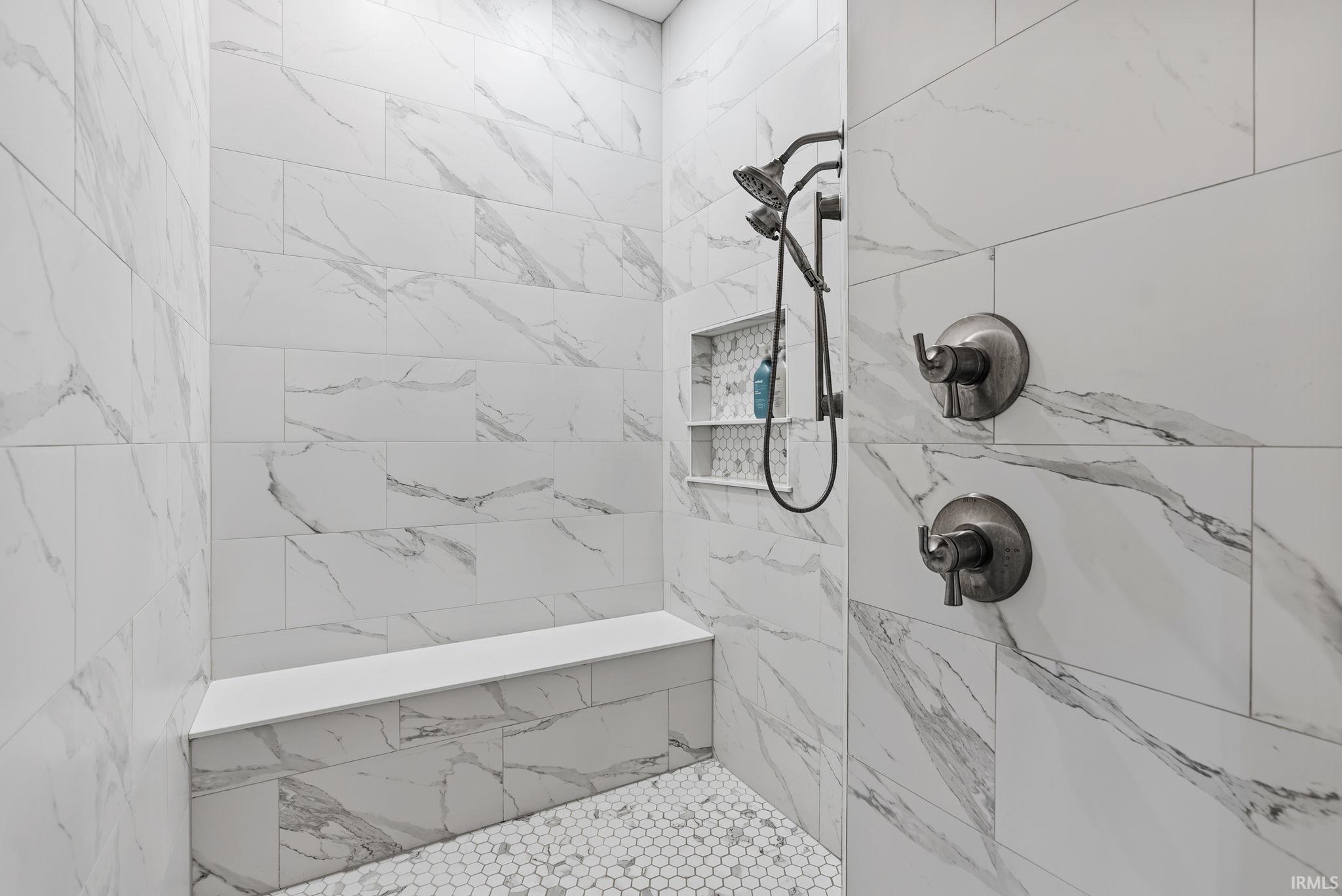 Walk-in shower featuring large format white tile with gray veining, a built-in bench, hexagon mosaic floor tile, a recessed niche with shelving, and dark-toned shower fixtures