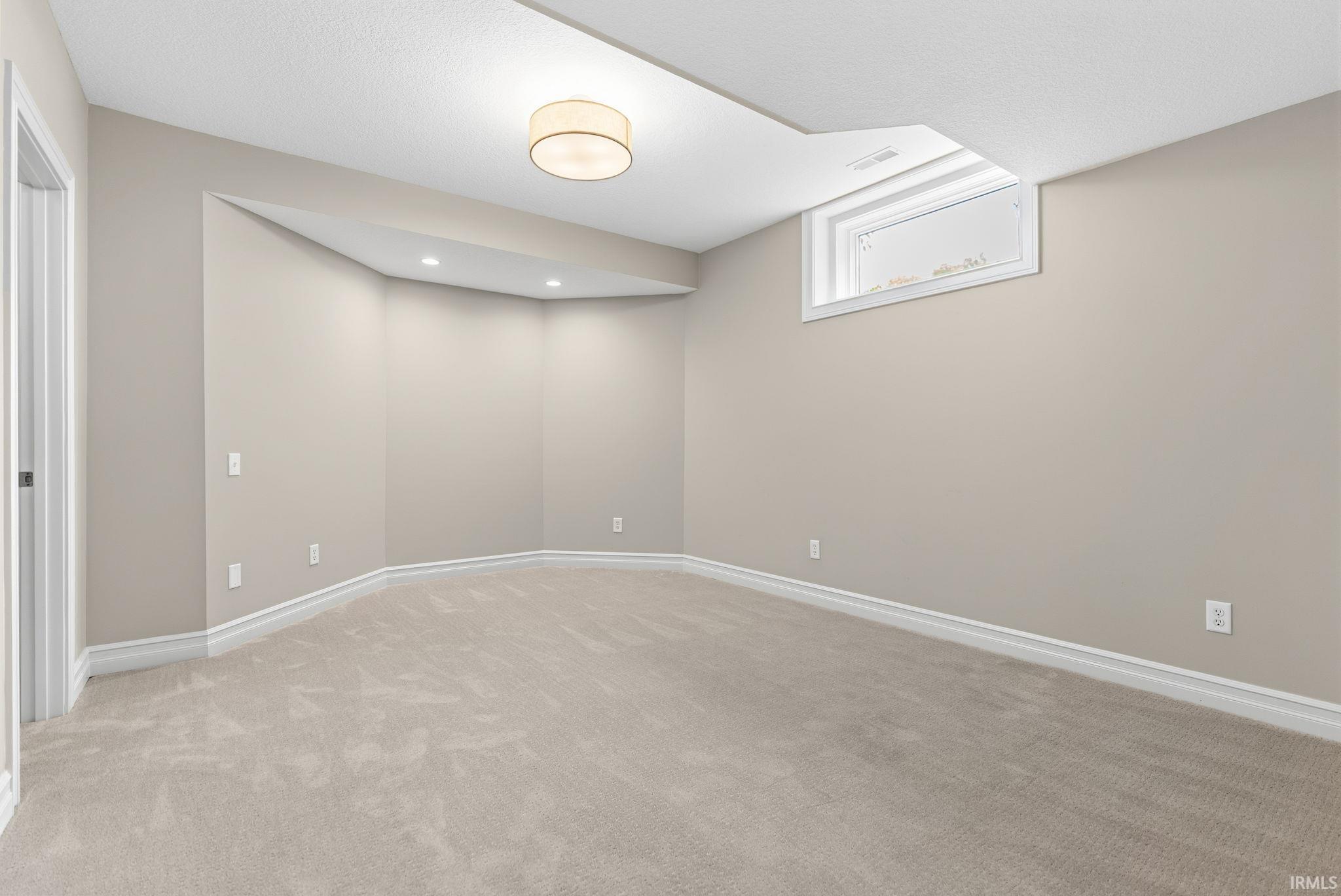 Finished lower-level space featuring neutral-toned carpeting and walls, a recessed ceiling section with integrated lighting, a ceiling-mounted drum light fixture, and a white-trimmed window
