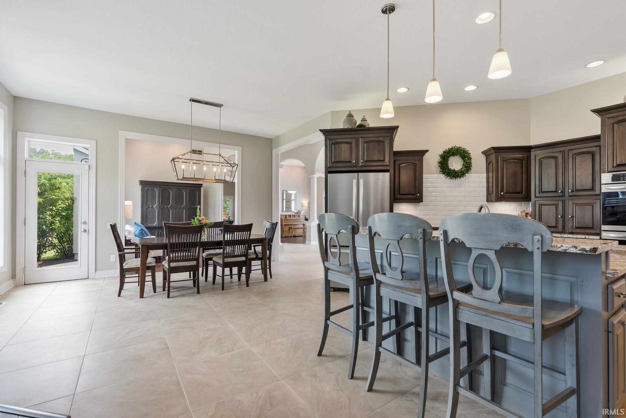 Open-concept kitchen and dining area featuring tile flooring, dark wood cabinetry, a subway tile backsplash, stainless steel appliances, and a kitchen island with pendant lighting