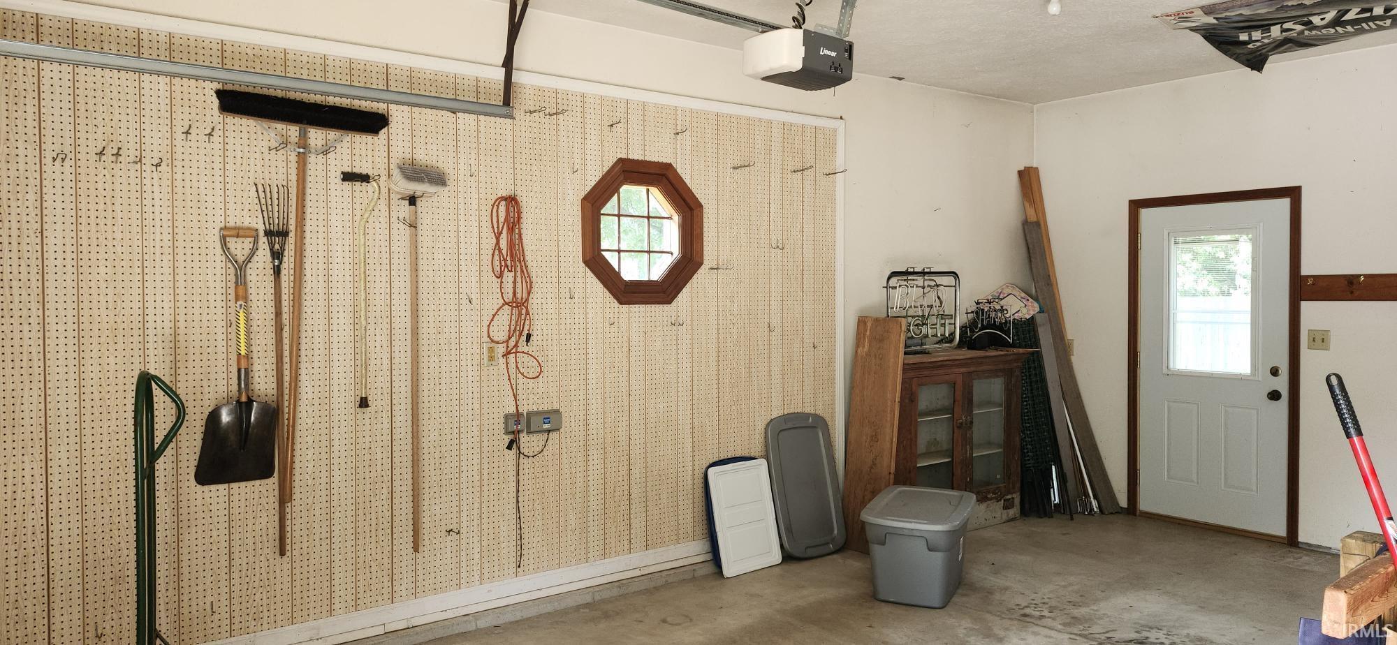 Attached garage featuring a concrete floor, light-toned pegboard wall, overhead garage door opener, octagonal window with wood trim, and a pedestrian door with an external light source