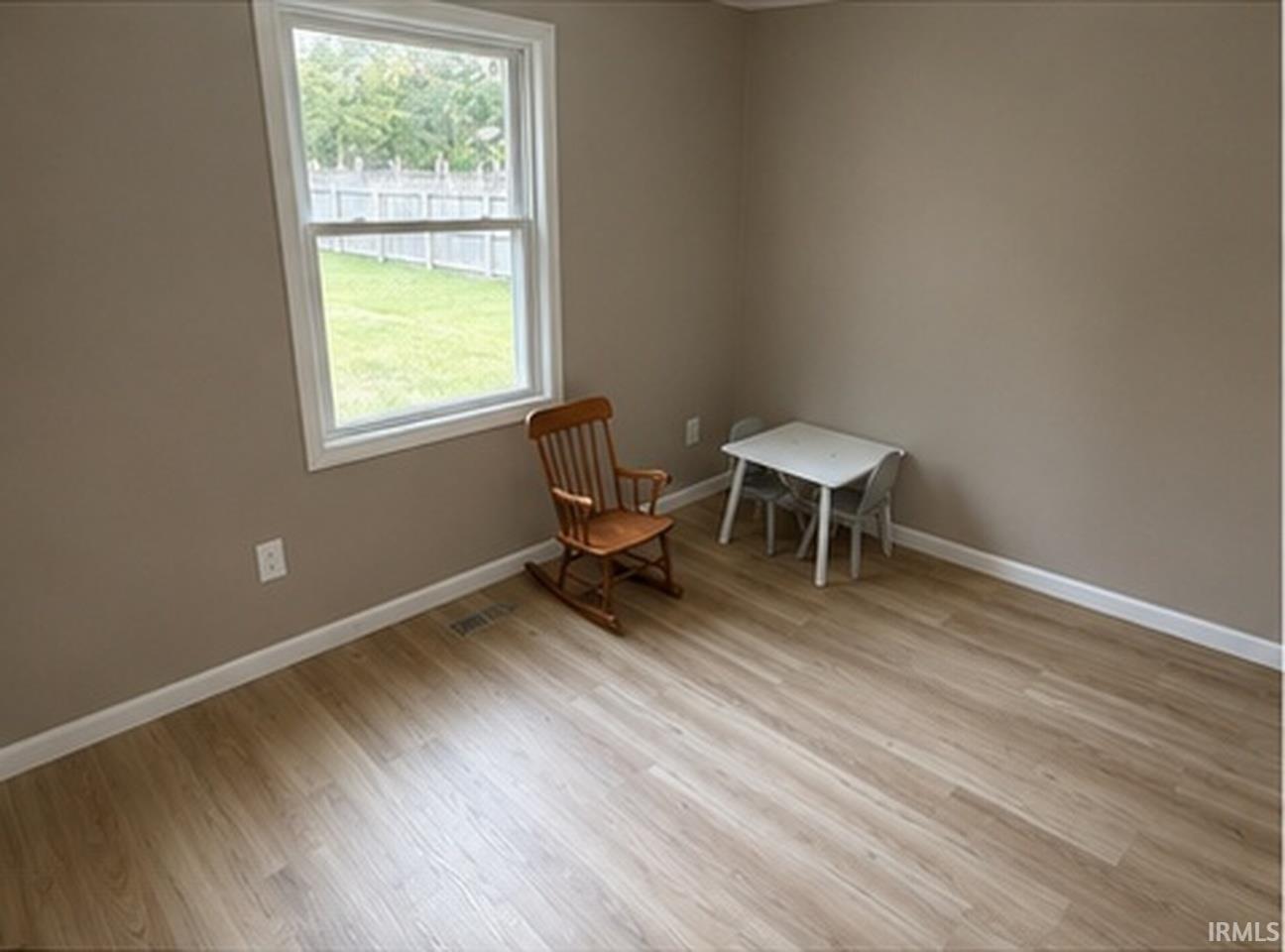 Neutral tone wall paint, wood-finish flooring, white window trim, white baseboards, and a backyard area with green space and a privacy fence