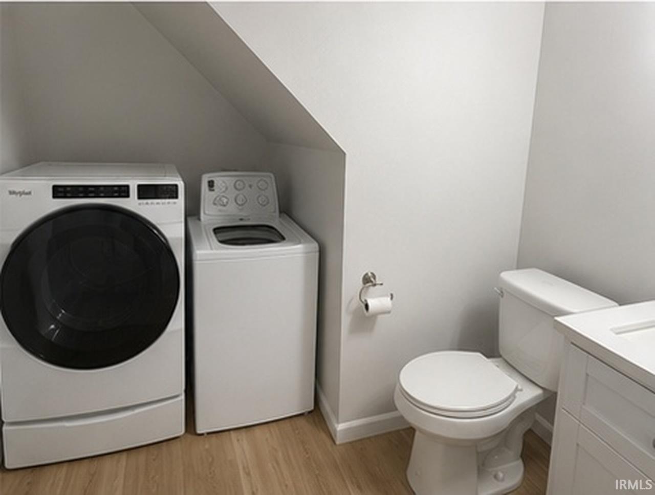 Compact laundry area with a white front-load washer and a top-load washing machine, complemented by a white toilet, toilet paper holder, and a white vanity with integrated sink