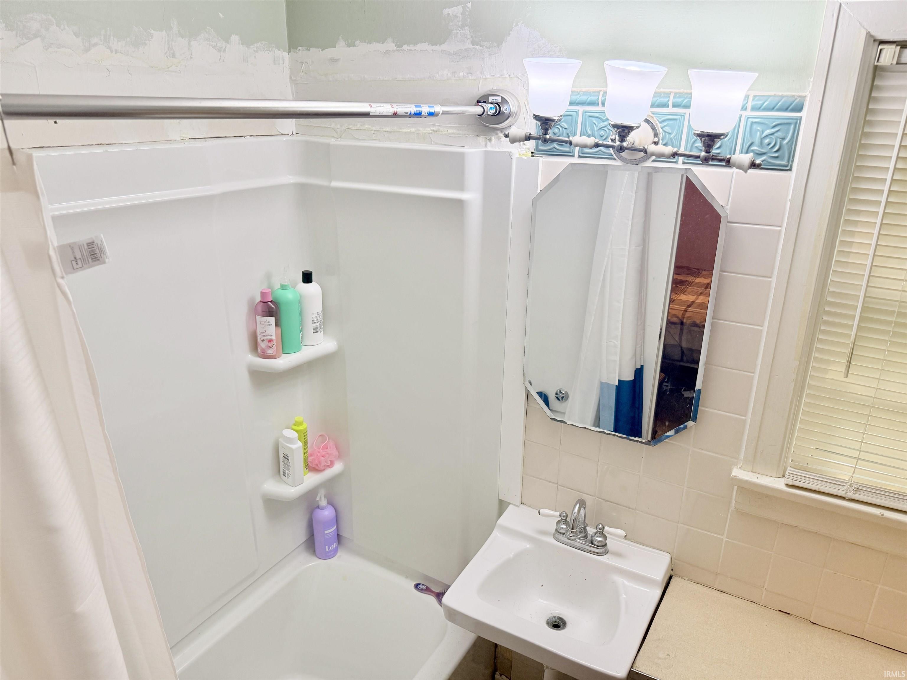 Bathroom featuring a white tub-shower combination with built-in corner shelving, a wall-mounted sink, an octagonal mirror, and a three-light vanity fixture