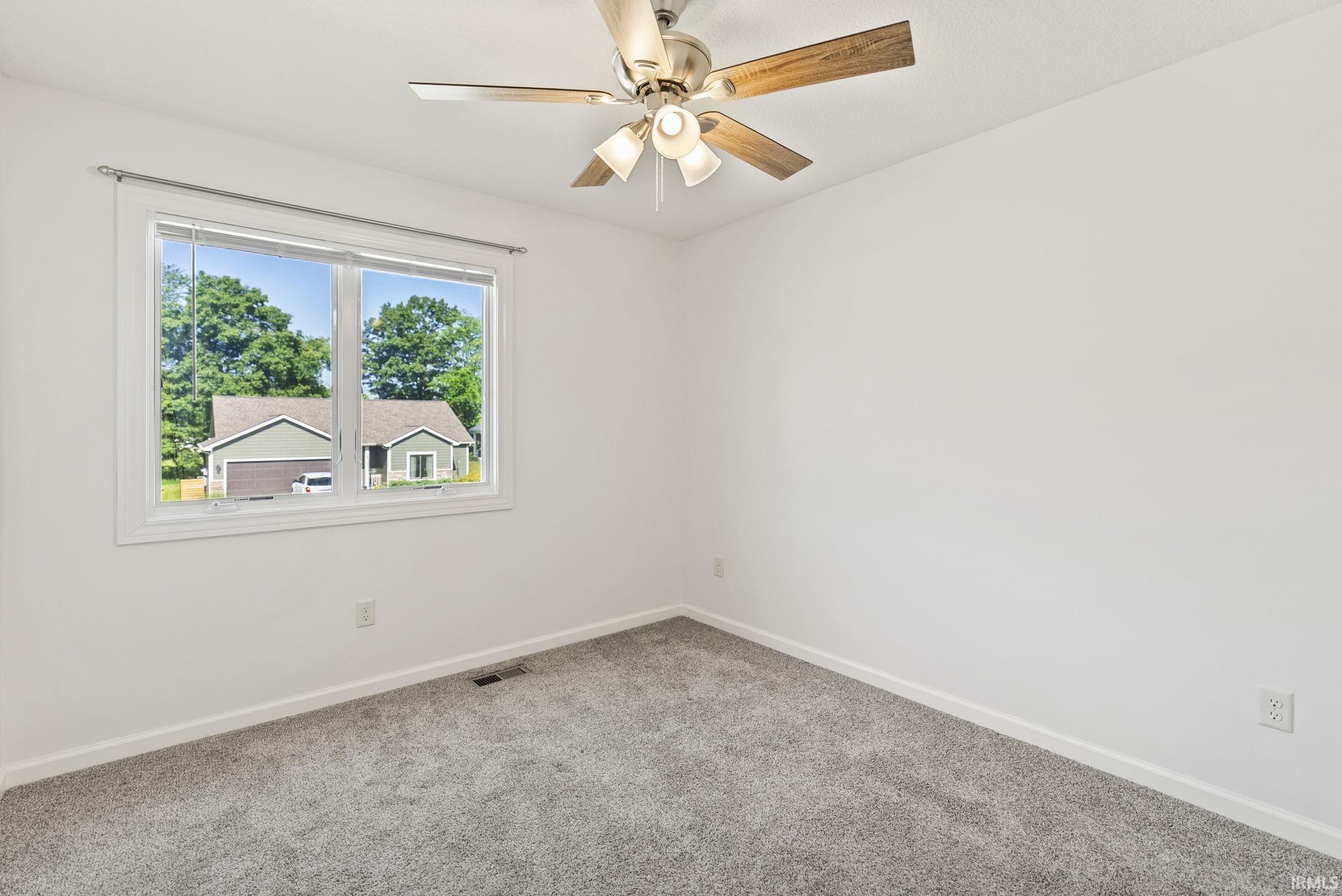 Bright interior space featuring a window with white trim, gray carpet, white walls, white baseboards, and a ceiling fan with wood-finish blades