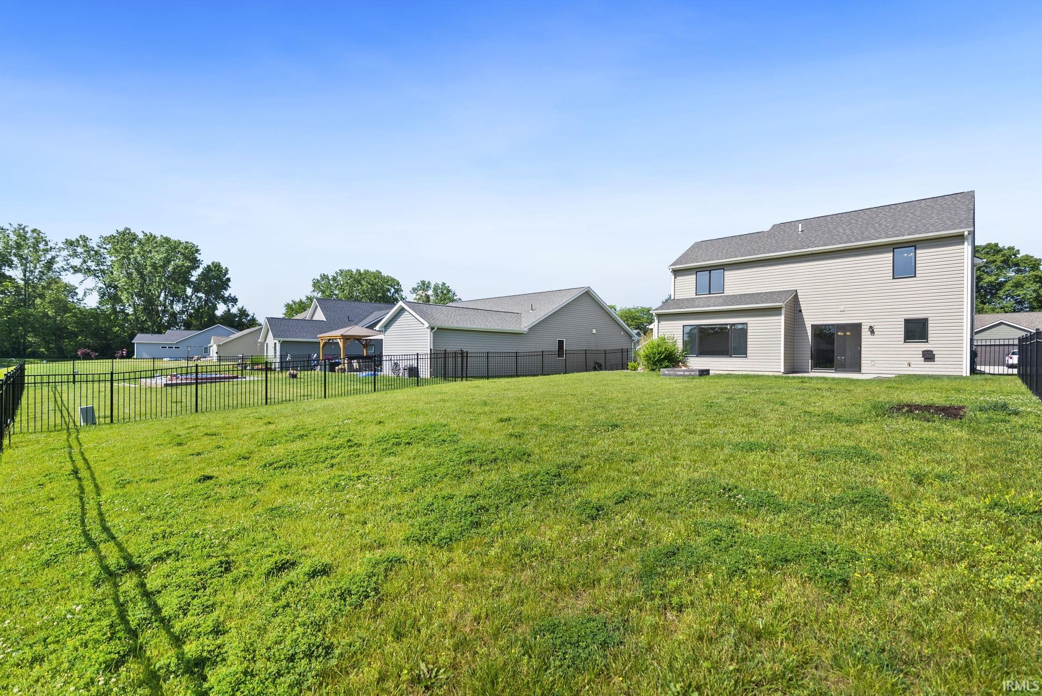 Expansive grassy backyard featuring a black metal fence, a two-story residence with light-colored siding, multiple windows, and a sliding glass door