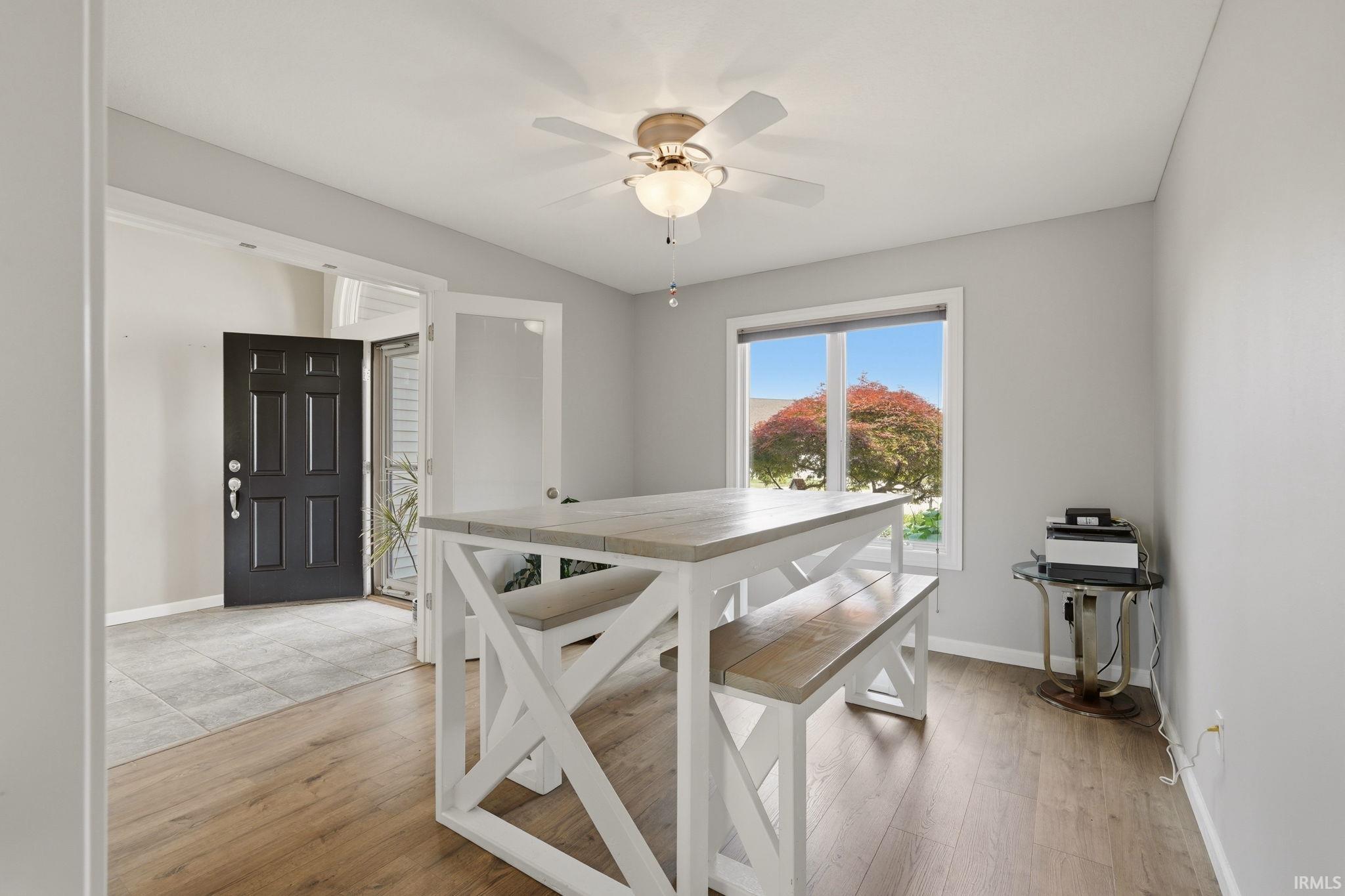 Wood-finish flooring throughout, featuring a window with exterior tree scenery, a ceiling fan with integrated lighting, and a doorway leading to an entryway with tiled flooring and a dark paneled door
