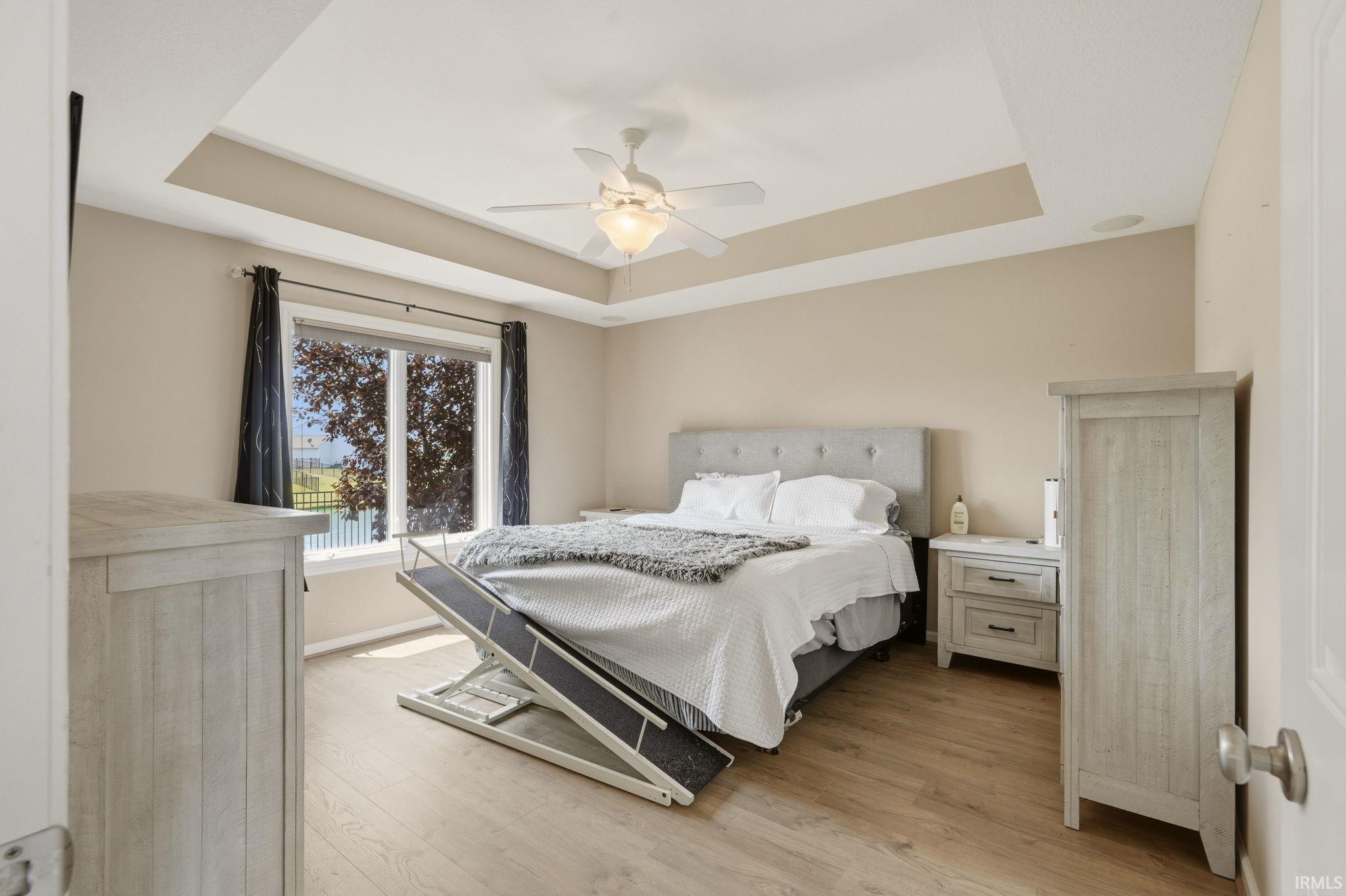 Recessed tray ceiling with central fan light, large window, and wood-finish flooring