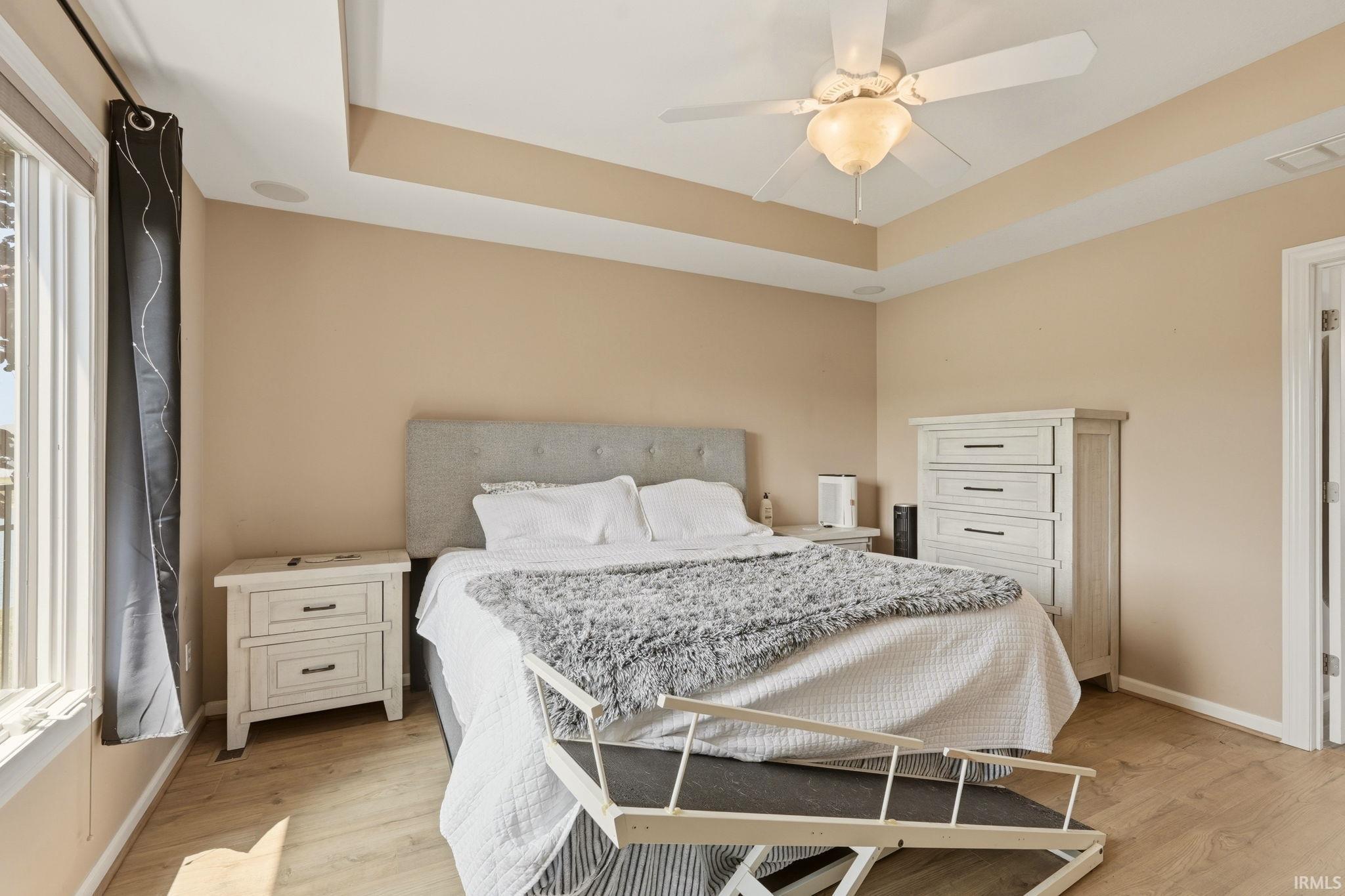 Spacious bedroom featuring light wood-finish flooring, a tray ceiling with a ceiling fan, and large windows with dark curtains