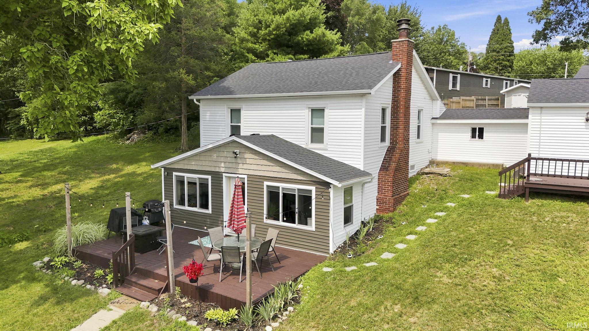 White siding residence featuring a large rear deck, a brick chimney, a detached garage with white siding, and a sloped grass yard