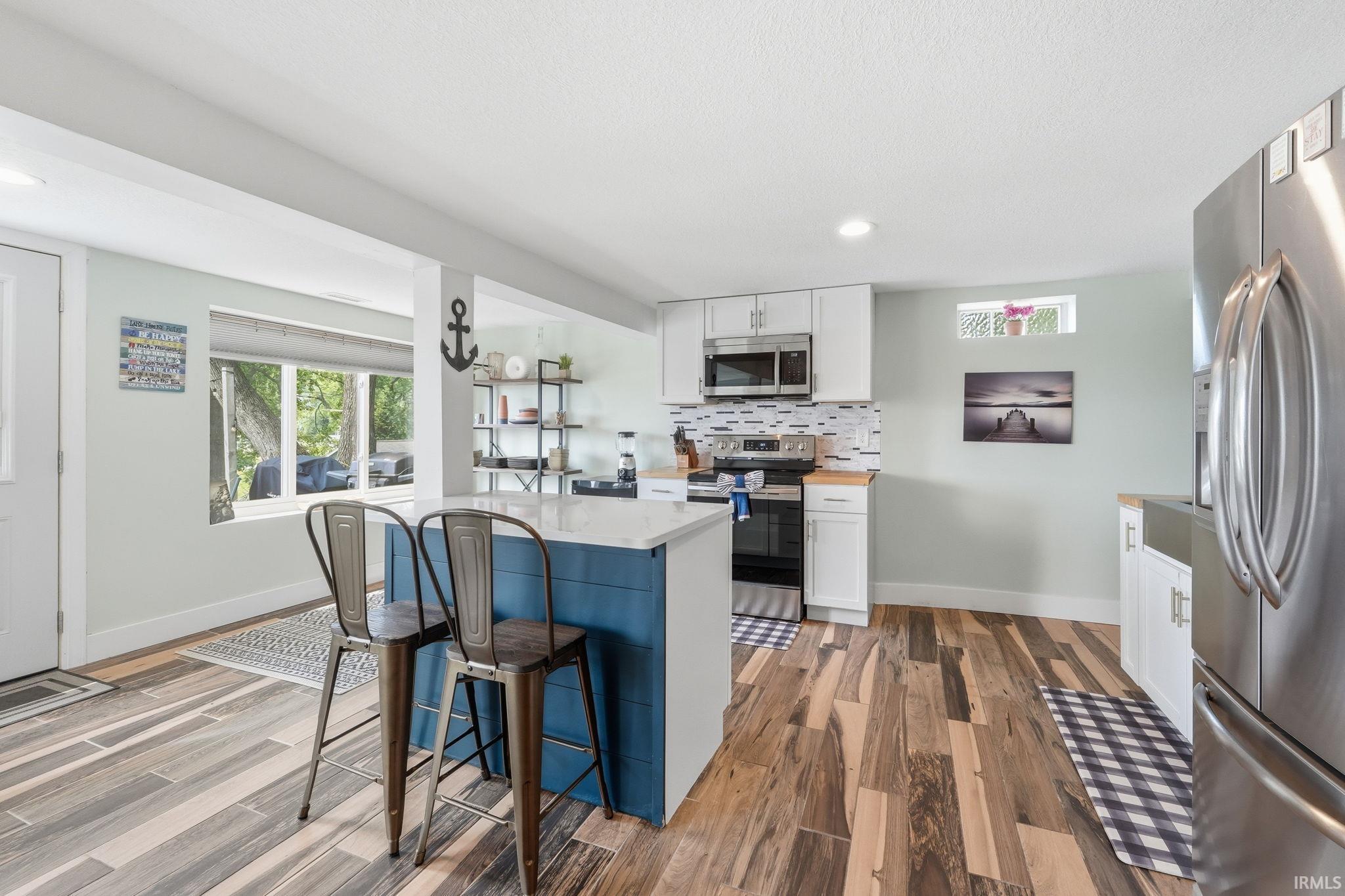 Modern kitchen featuring a blue island with a light-toned countertop, stainless steel appliances, white cabinetry, a patterned tile backsplash, and multi-toned wood-finish flooring