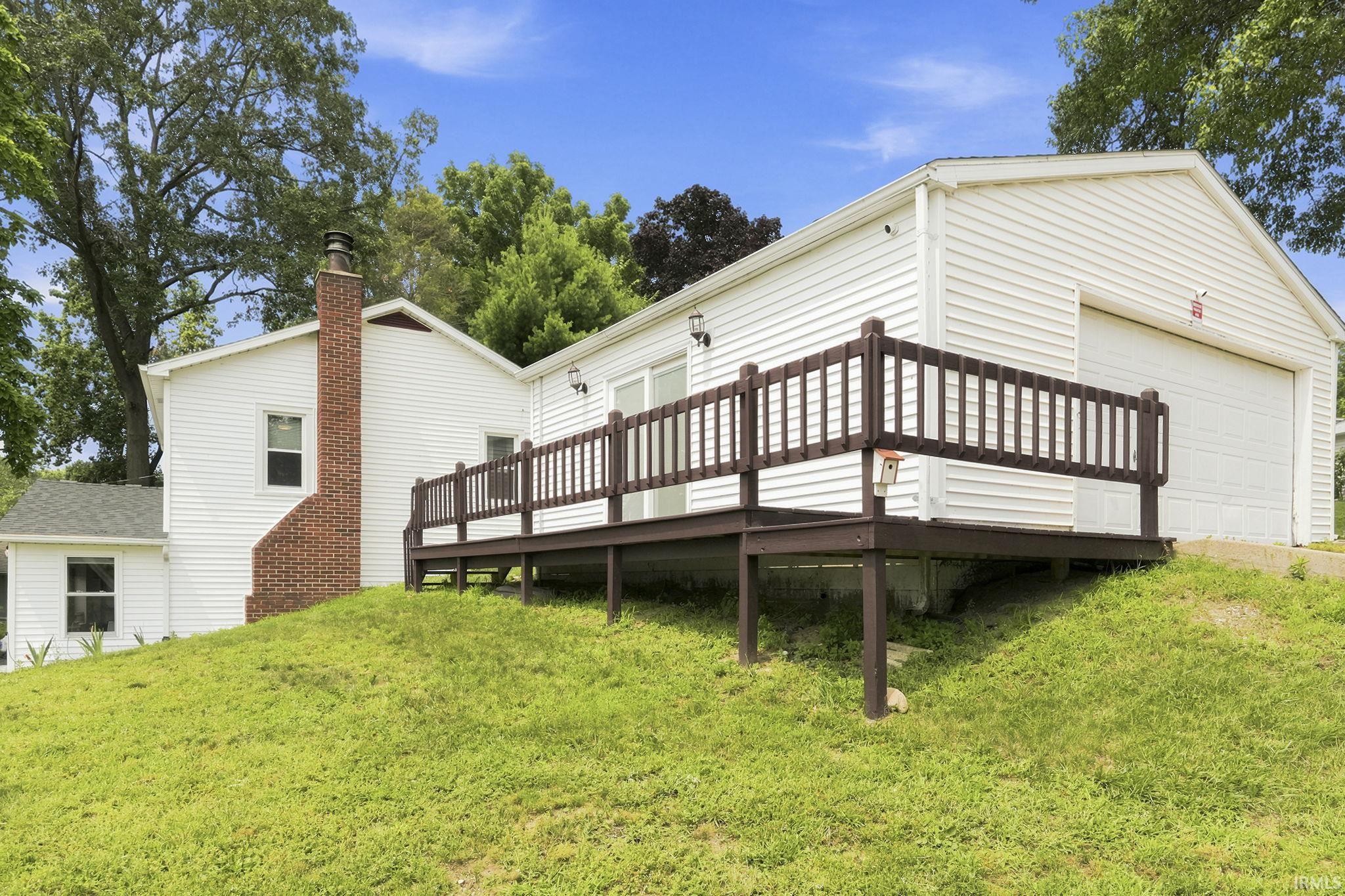Exterior featuring a white siding finish, a prominent brick chimney, a dark-stained wood deck with railings, an attached garage with a white door, and surrounding mature trees