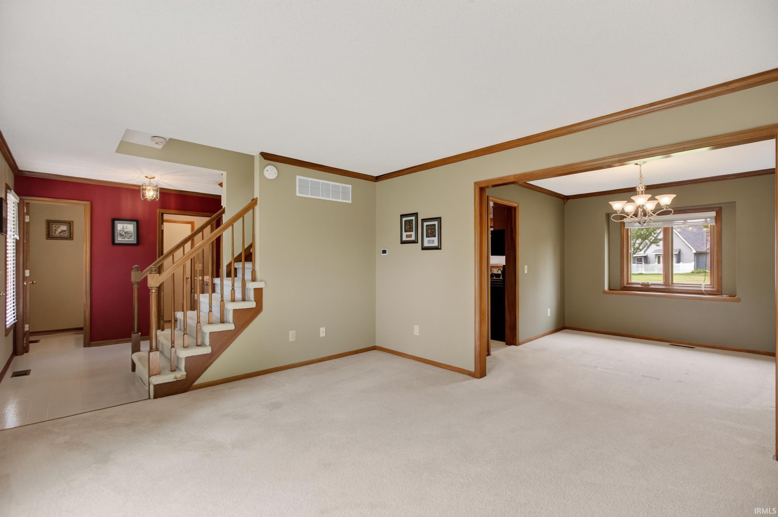 Carpeted living area with wood trim and entryway, featuring a staircase with wood banister and carpeted treads, a framed opening to an adjacent room with a chandelier, and a window with wood framing