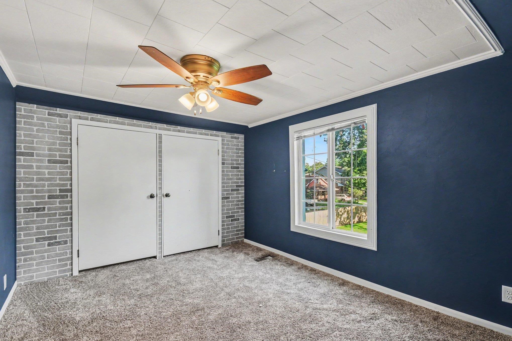 Textured ceiling panels, ceiling fan with integrated lighting, double bi-fold closet doors, accent wall with brick-finish paneling, and a white-framed window