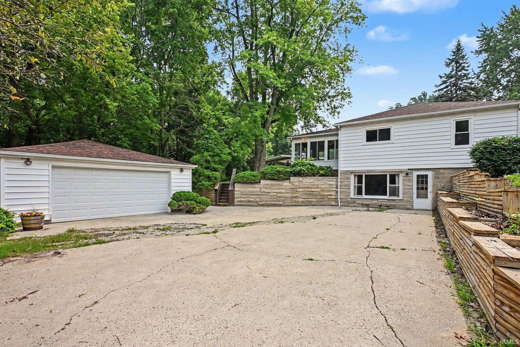 Exterior featuring white siding, a two-car garage with a brown shingle roof, and a stone retaining wall