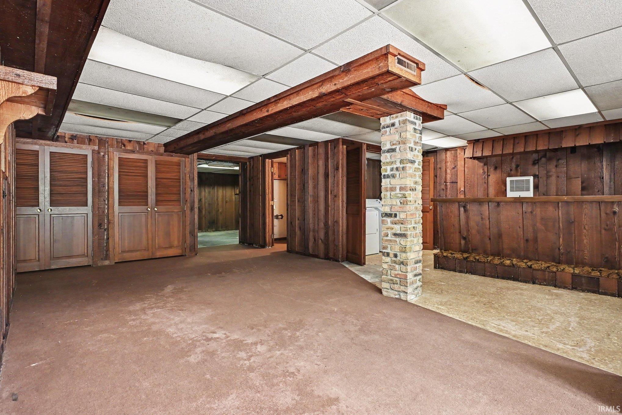 Finished basement featuring wood paneling, exposed wood beams, a brick pillar, drop ceiling with integrated lighting, and multiple storage closets