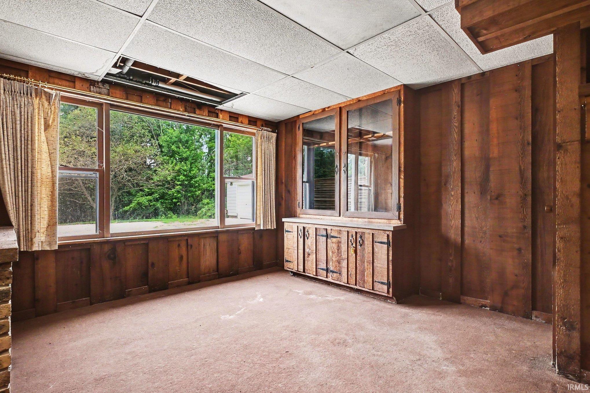 Room featuring wood-paneled walls, a large picture window, and a built-in wooden cabinet with glass-fronted upper storage