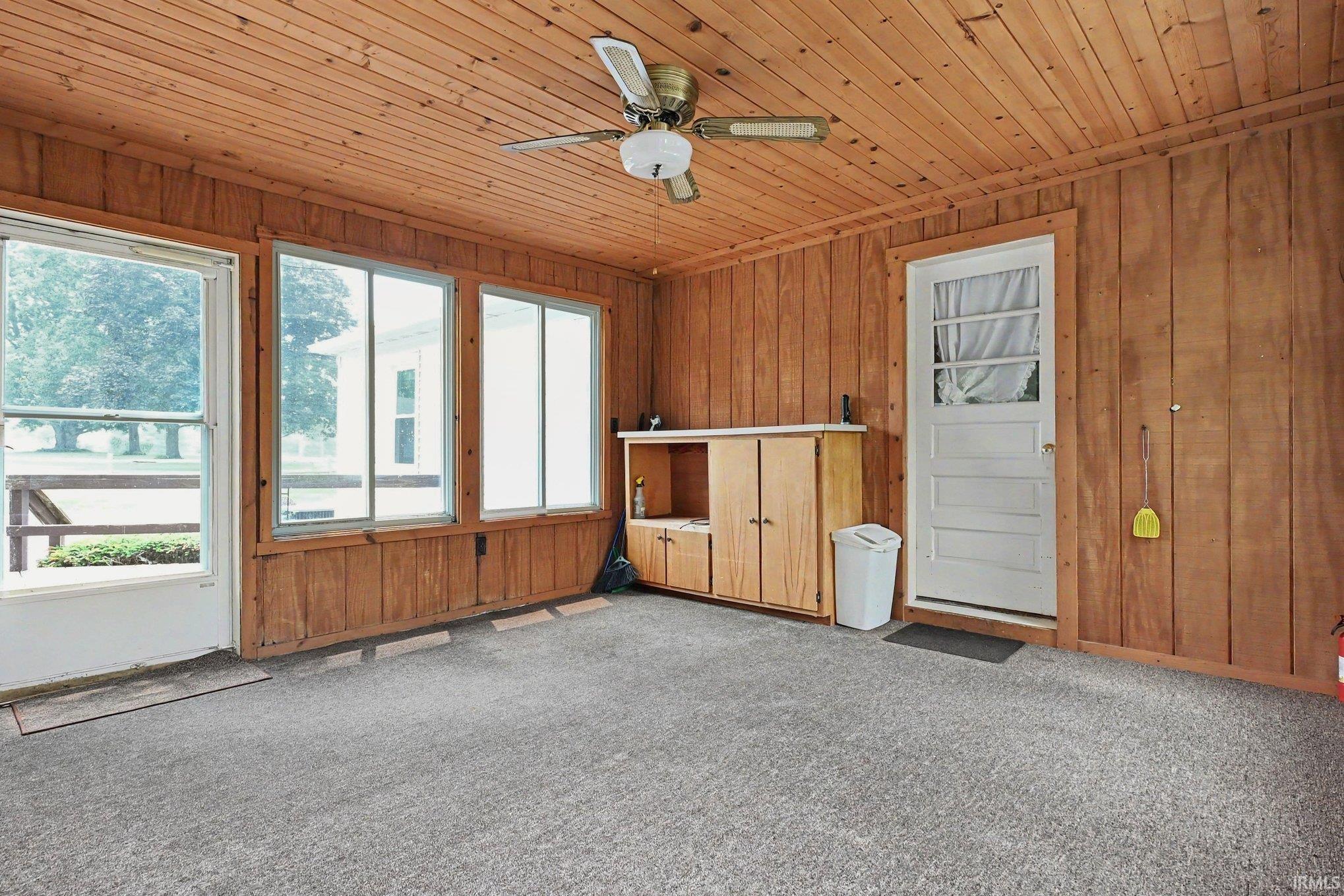 Room featuring wood paneling on walls and ceiling, grey carpet flooring, multiple windows, a ceiling fan, and built-in cabinetry
