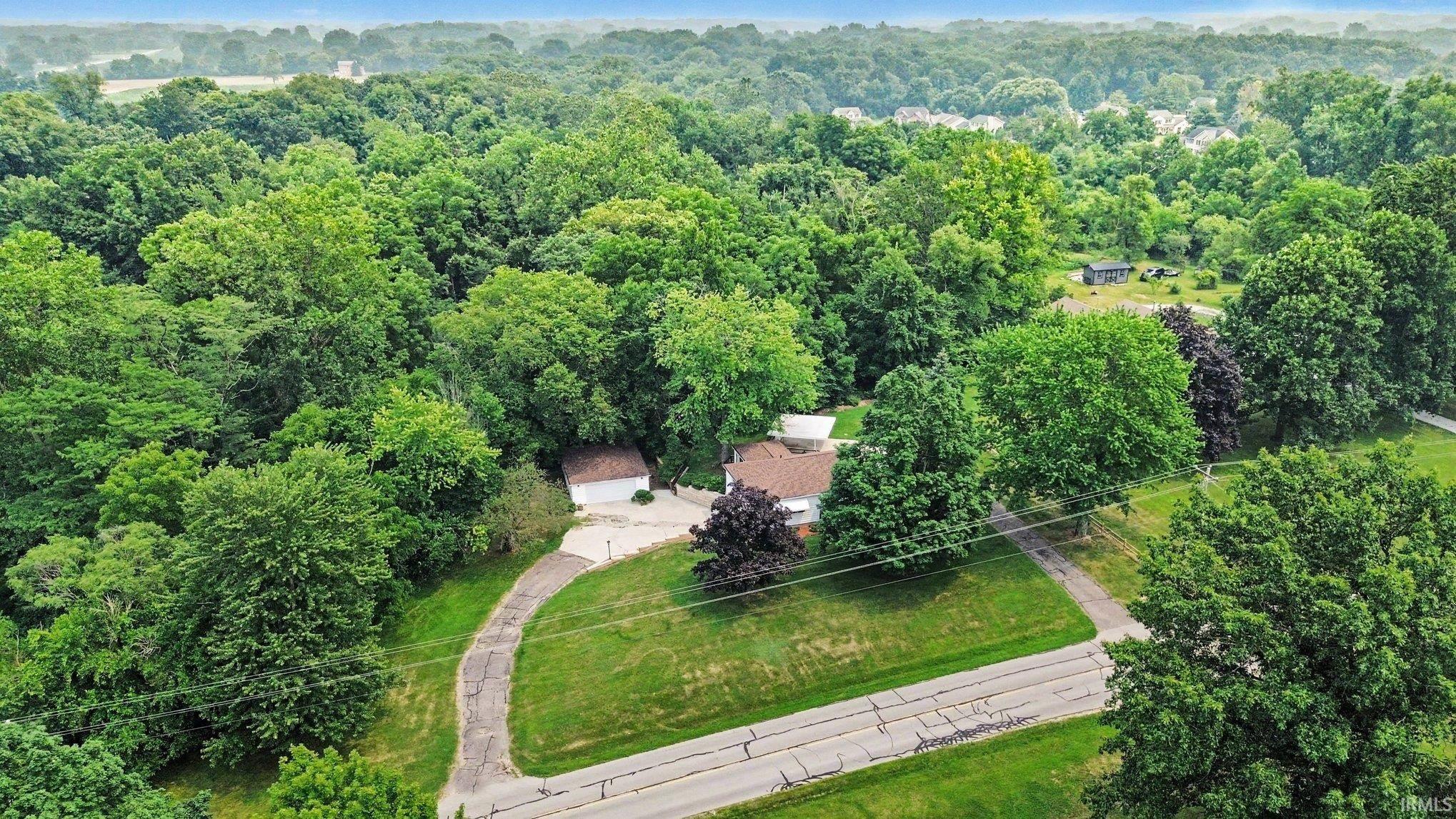 Expansive tree canopy surrounding the property, featuring a private driveway, manicured lawn, and a light-colored exterior residence with a brown roof