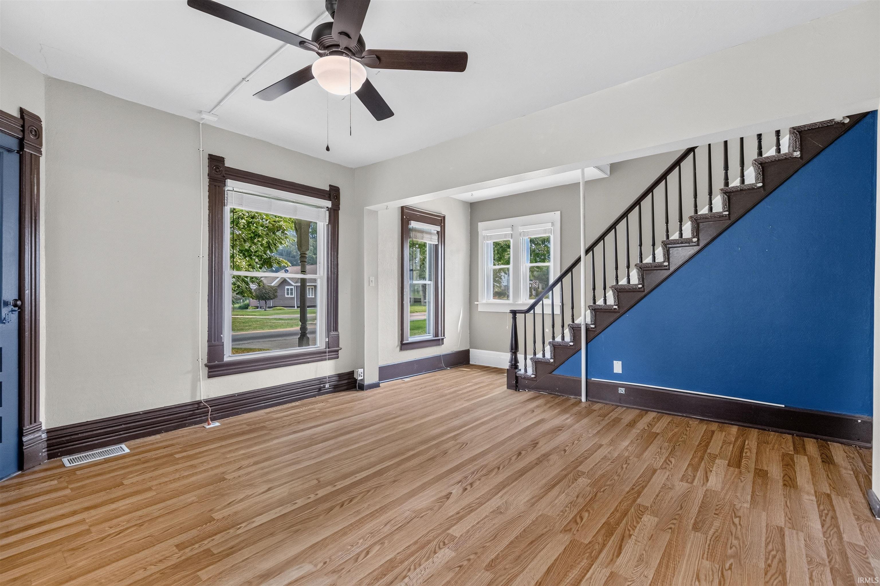 Expansive open room featuring wood-finish flooring, a ceiling fan with integrated lighting, and a staircase with dark wood treads and a contrasting blue wall