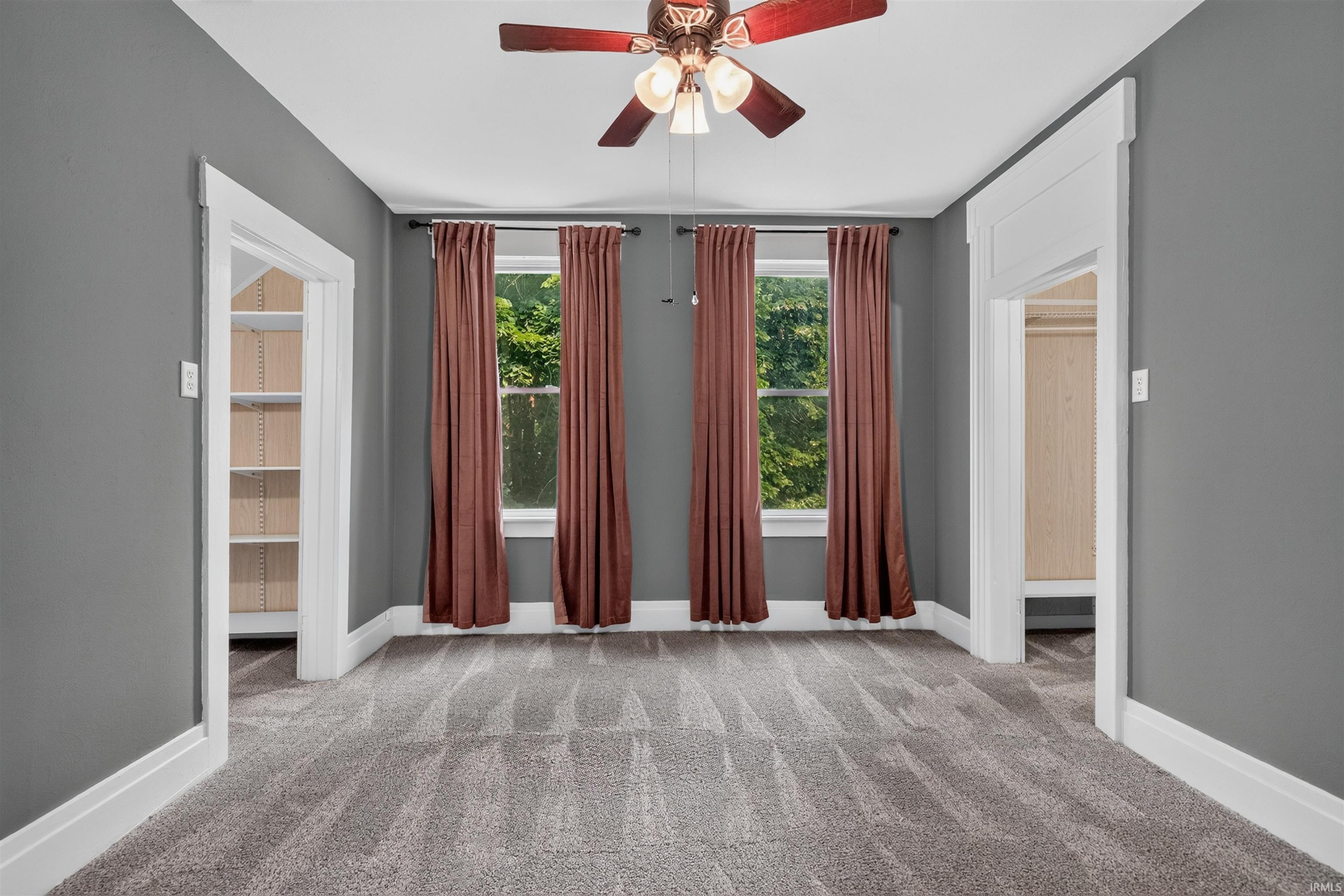 Room featuring plush gray carpet, two windows with brown drapery, a ceiling fan with integrated lighting, and two doorways framed with white trim