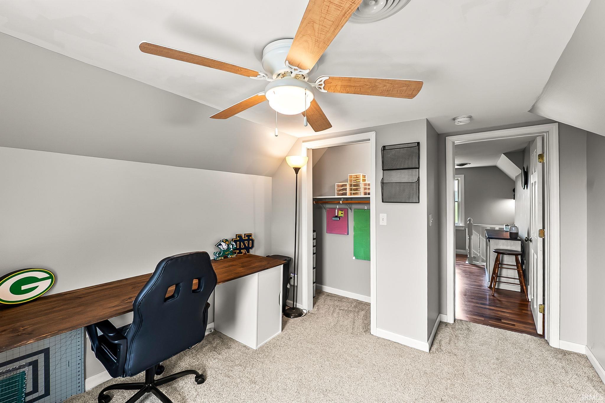 Carpeted room featuring an angled ceiling, wood-finish ceiling fan with integrated lighting, and a built-in wood-finish desk spanning the wall