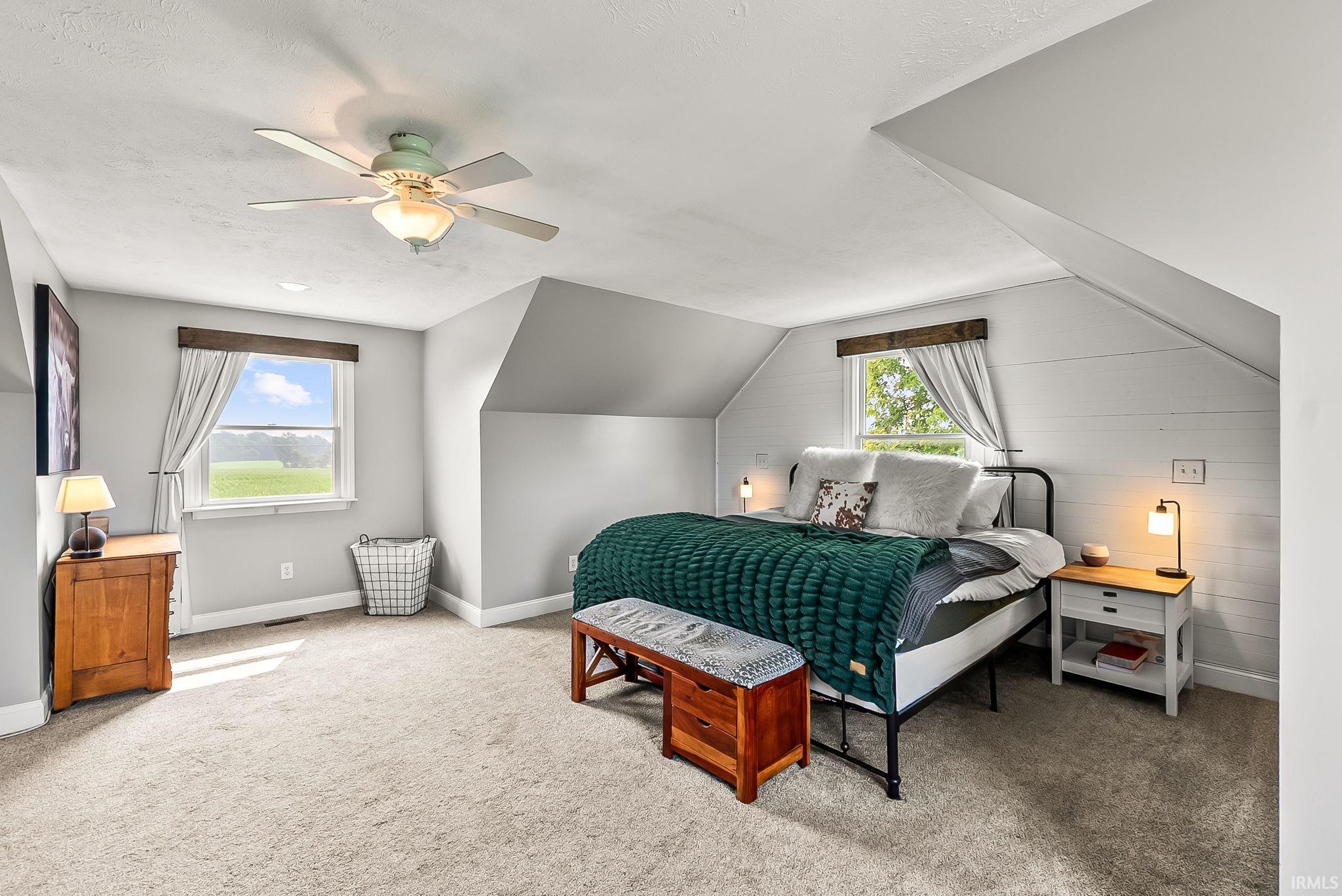 Carpeted room featuring a ceiling fan with light, two windows with wood-finish trim, a sloped ceiling, and white shiplap accent wall