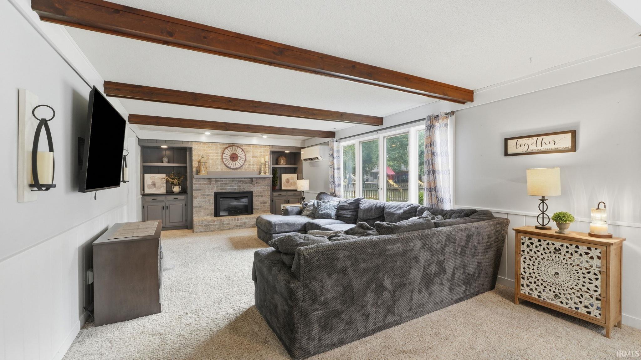 Living area featuring exposed wood-finish ceiling beams, an integrated brick fireplace, built-in shelving, white wainscoting, and large windows
