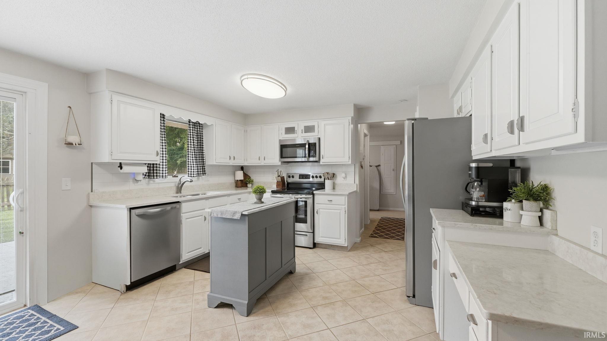 Bright kitchen featuring white cabinetry, light-toned countertops, stainless steel appliances, a center island with storage, and neutral tile flooring