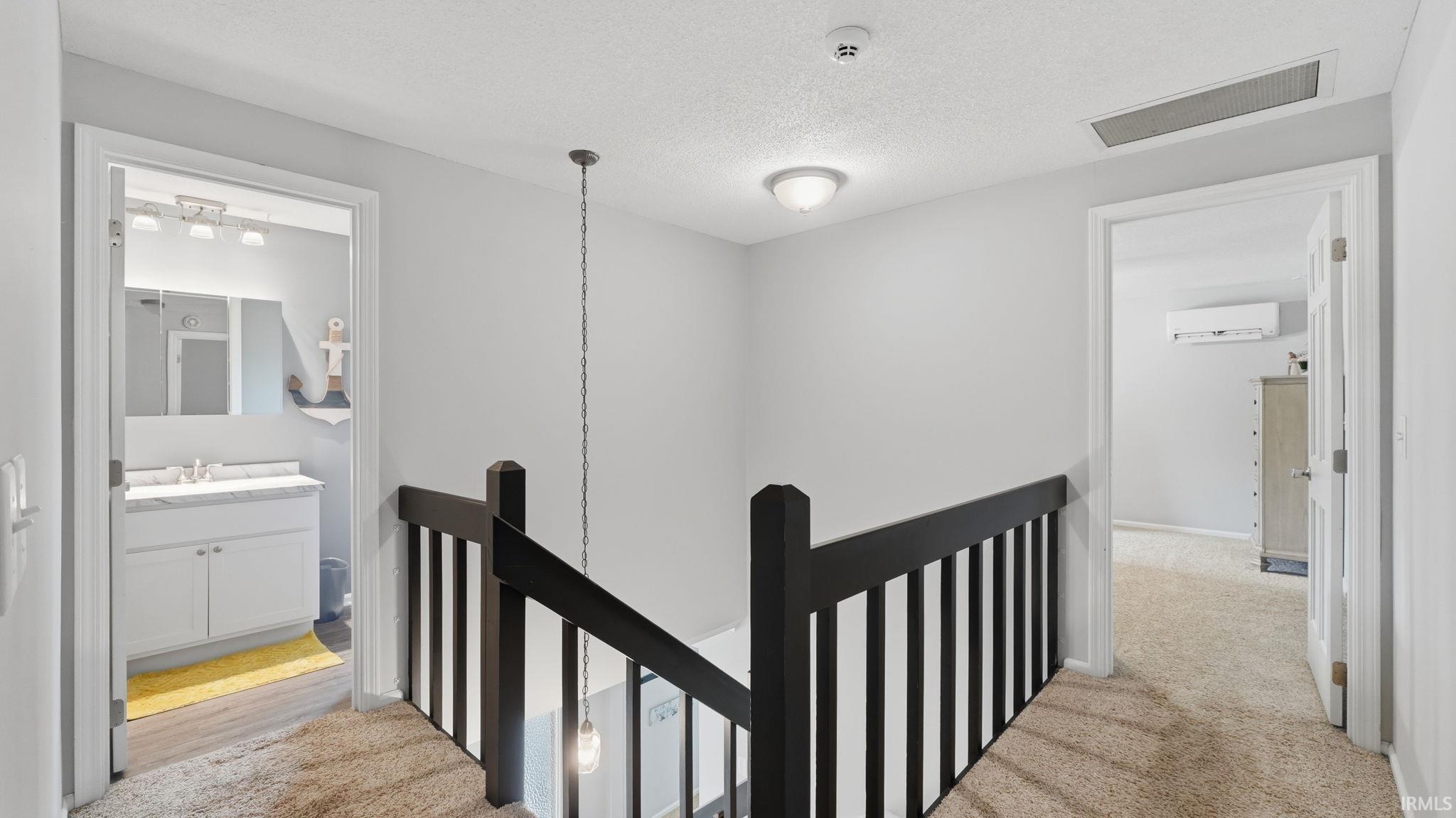 Upper-level landing featuring neutral carpeting, a dark wood-finish staircase railing, and access to a bathroom with a white vanity and a separate room with an installed wall-mounted unit