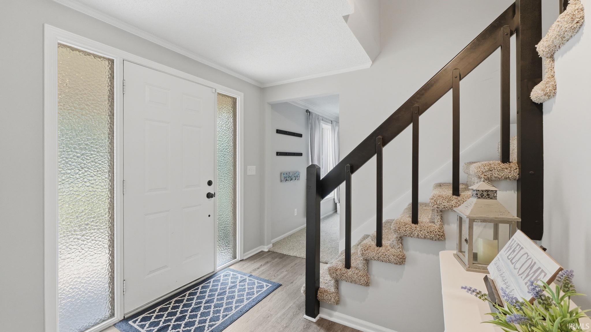 Foyer entrance featuring a white panel door with textured side lights, wood-finish flooring, and a carpeted staircase with dark wood railings