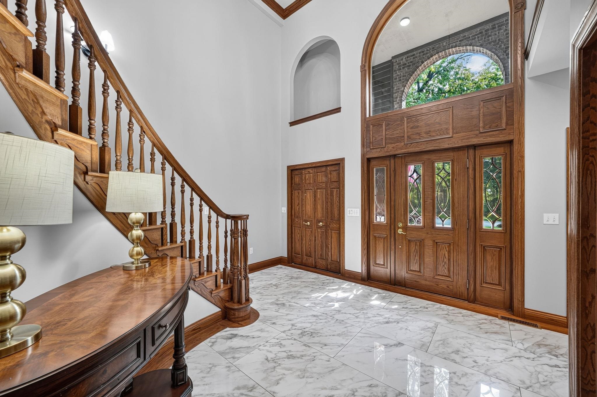 Grand entryway featuring a sweeping hardwood staircase, marble-finish tile flooring, double-door entry with decorative glass inserts, and a prominent arched transom window