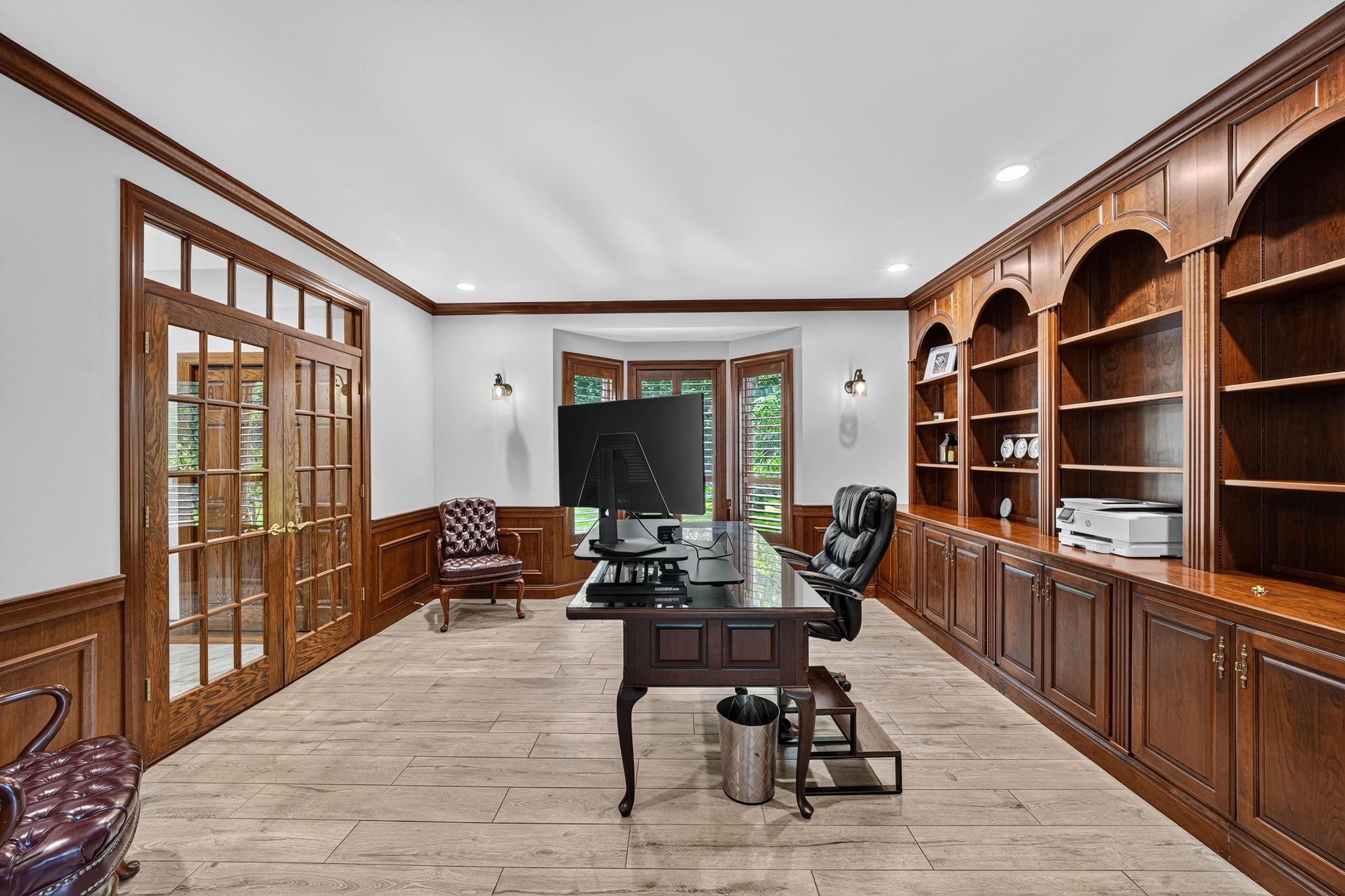 Wood-paneled office featuring extensive built-in shelving, glass-paned double doors, a bay window, recessed lighting, and wood-finish flooring