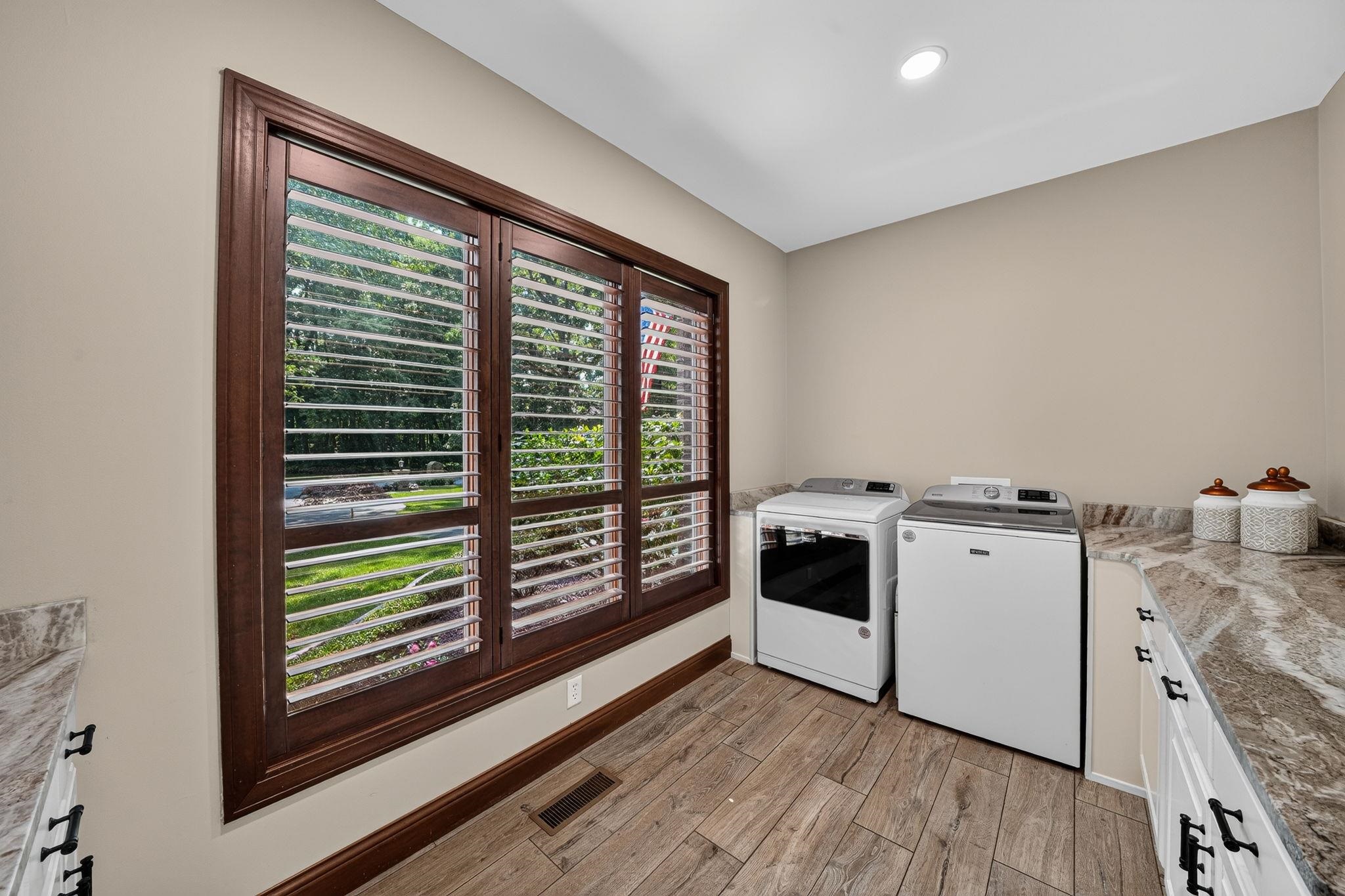 Utility room featuring wood-finish flooring, a large window with plantation shutters, a lengthy countertop, and white cabinetry with black hardware